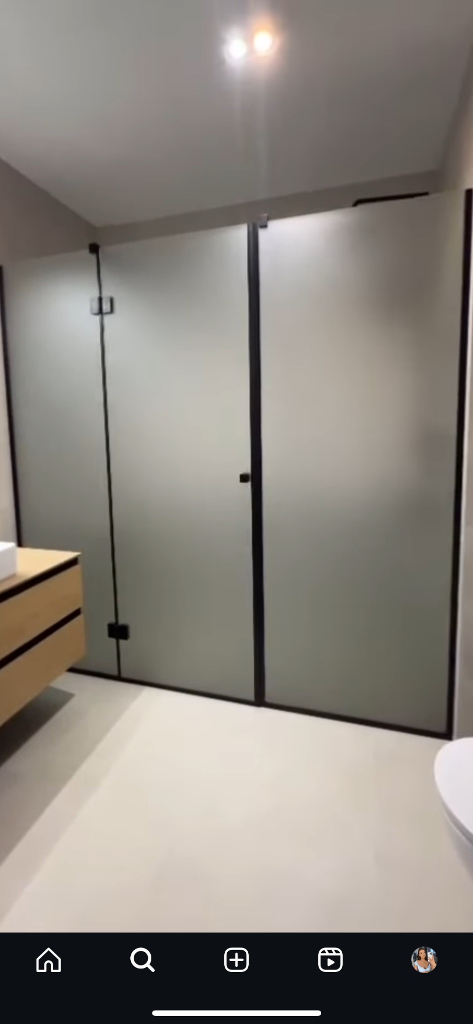 Matowe szklane drzwi prysznicowe z czarnymi ramami i zawiasami w nowoczesnej łazience, widok na umywalkę z drewnianymi szufladami i jasną podłogę.