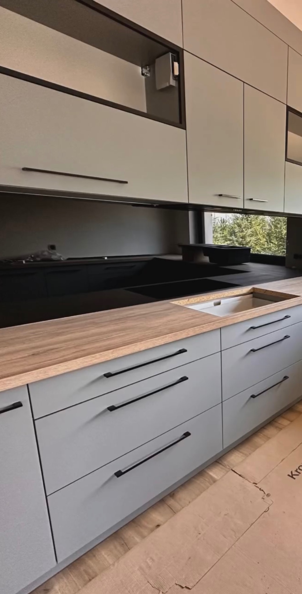 Szare szafki kuchenne z drewnianym blatem i czarnym szkłem na ścianie, widoczne przygotowane miejsce na zlew, podłoga zabezpieczona kartonem.