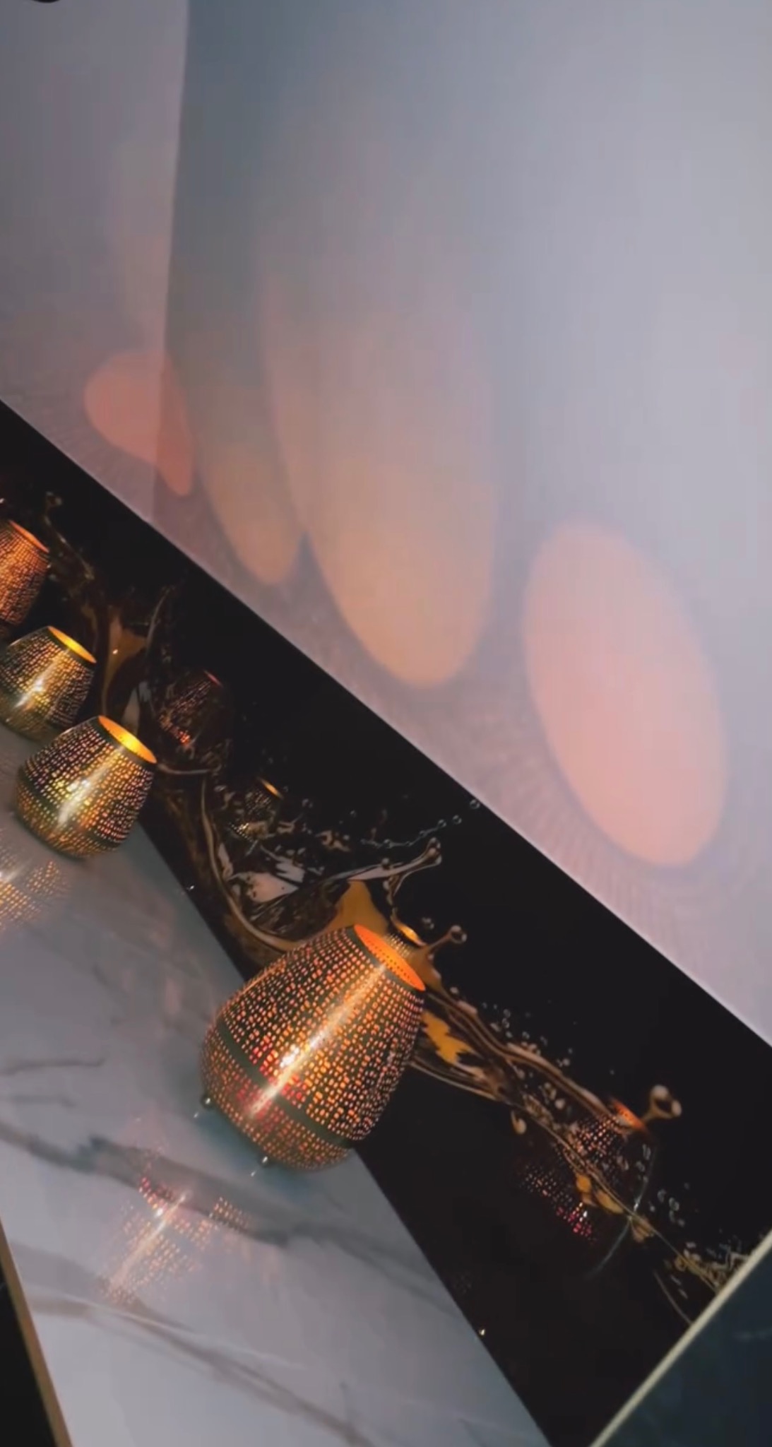 Dekoracyjny panel szklany z motywem rozbryzgującej się cieczy, oświetlony od wewnątrz ażurowymi lampionami, tworząc ciepłą, refleksyjną atmosferę.