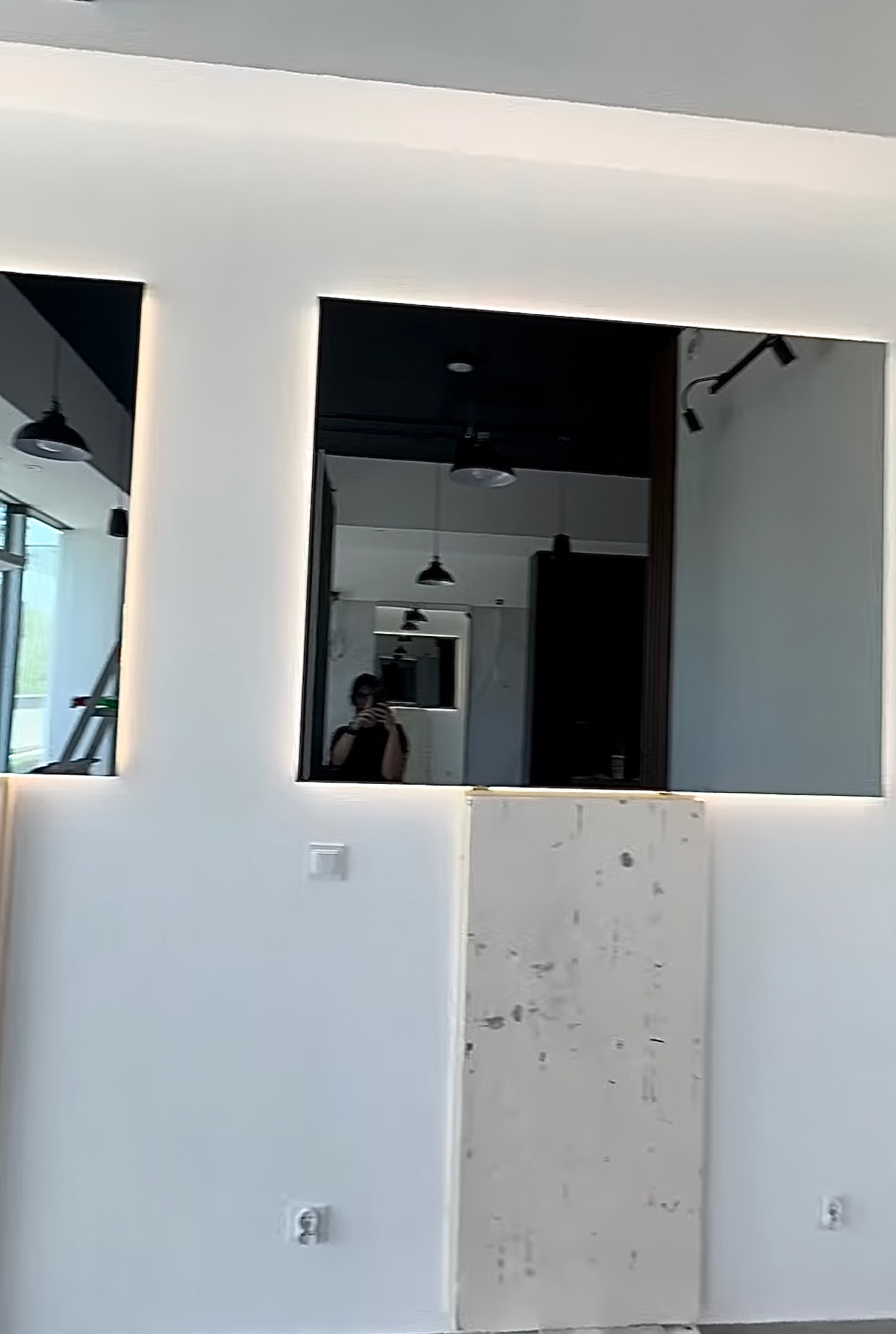 Dwa duże, prostokątne lustra z podświetleniem LED na białej ścianie, odbijające wnętrze pomieszczenia z widocznymi lampami sufitowymi i sylwetką osoby robiącej zdjęcie.