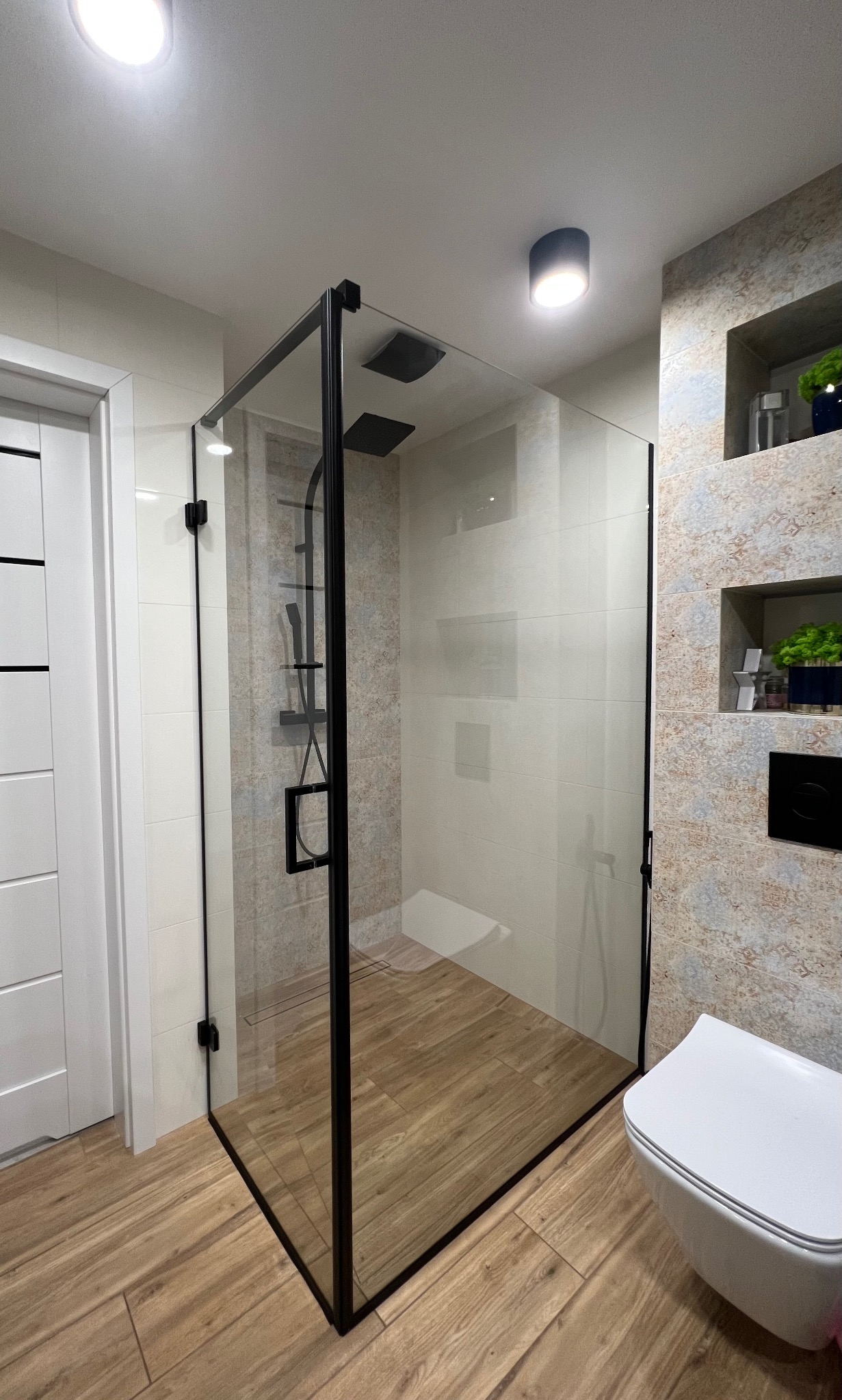 Nowoczesna kabina prysznicowa z czarnymi profilami, szklane ściany, drewnopodobna podłoga, deszczownica i bateria w czarnym macie, widok na fragment toalety.