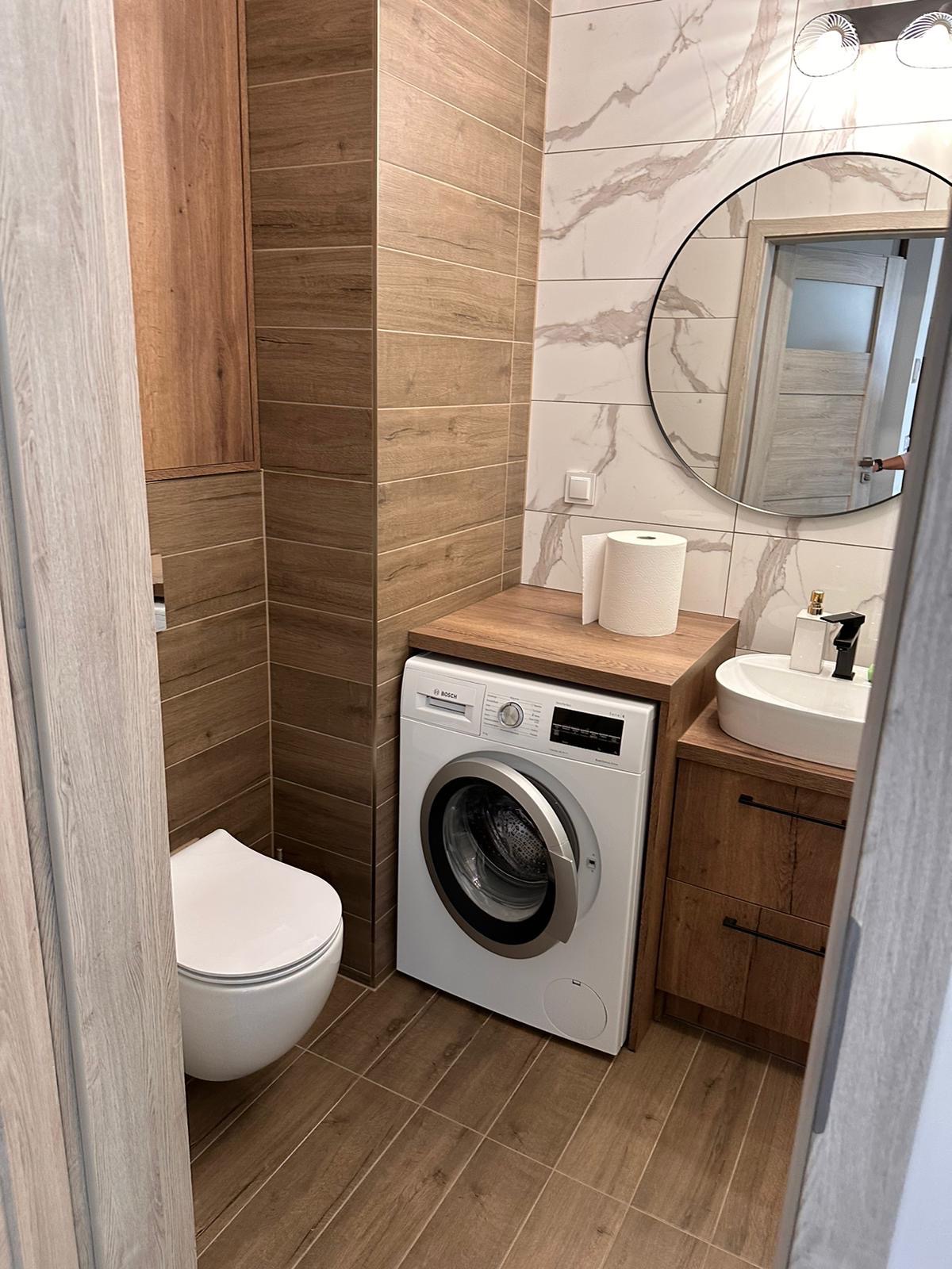 Wykończona łazienka z drewnopodobnymi płytkami, wiszącą toaletą, pralką pod blatem i okrągłym lustrem z podświetleniem.