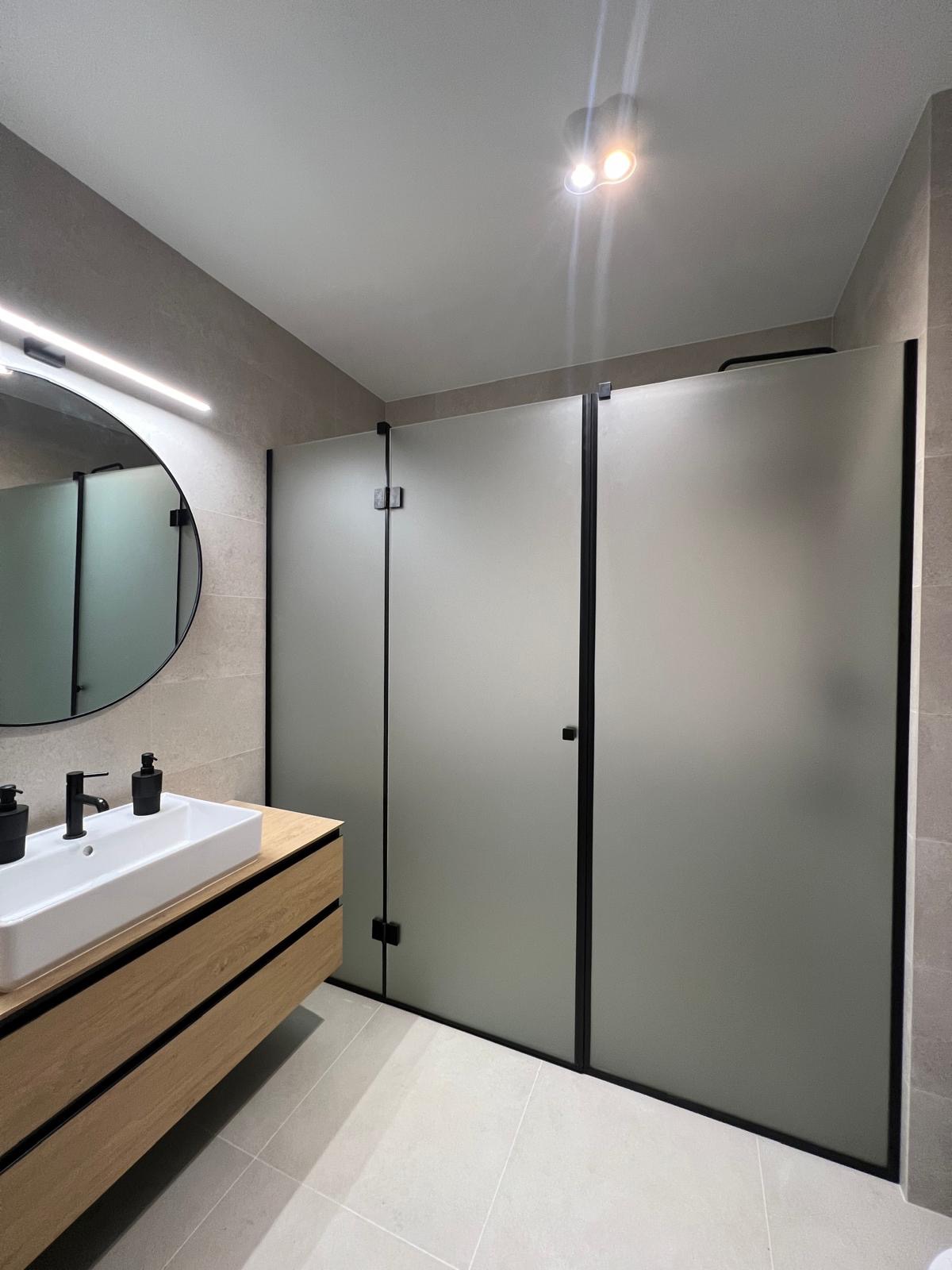 Nowoczesna łazienka z matowymi drzwiami kabiny prysznicowej w czarnej ramie, umywalką nablatową i okrągłym lustrem z podświetleniem LED.