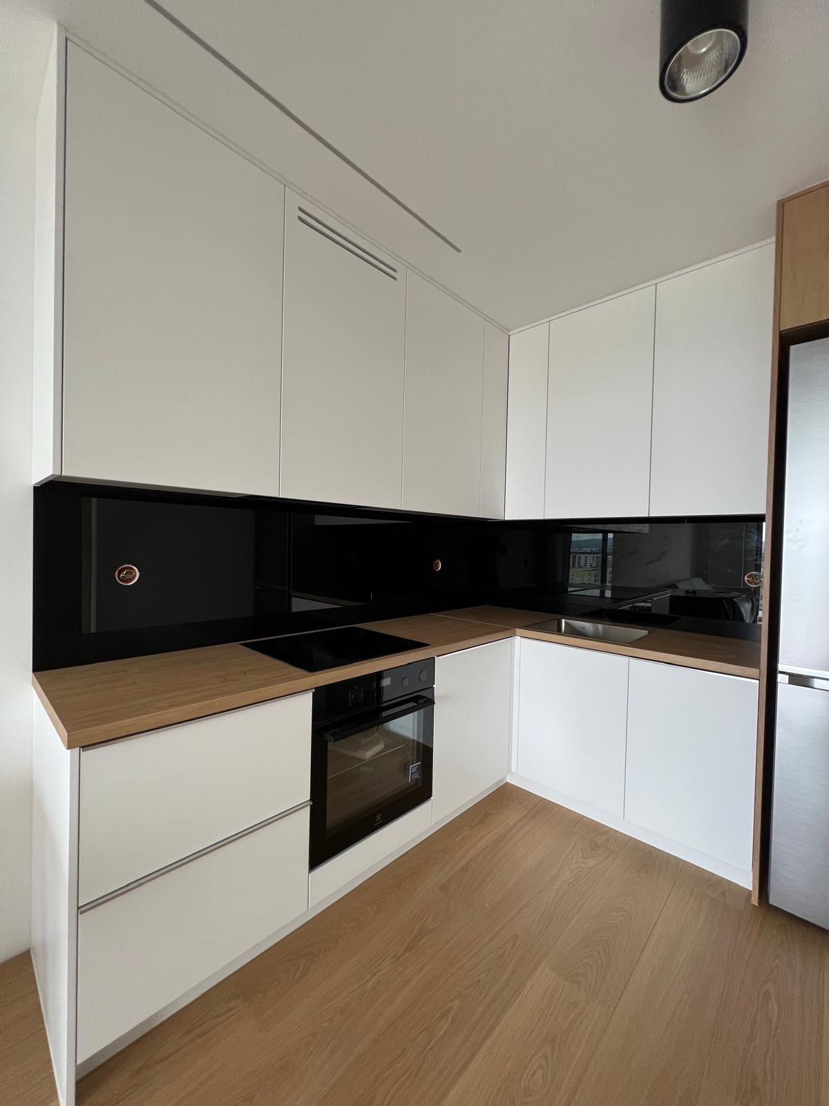 Nowoczesna kuchnia z białymi szafkami, drewnianym blatem i czarnym panelem szklanym na ścianie, widok na zabudowaną płytę indukcyjną i piekarnik.