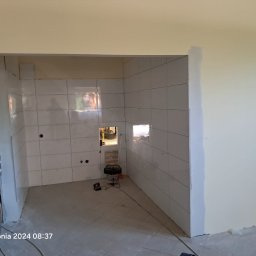 Michał Paszak - Wnętrze łazienki w trakcie prac wykończeniowych, widoczne białe płytki na ścianach, przygotowane otwory na instalacje, odkurzacz przemysłowy na podłodze, ściany częściowo pomalowane na kremowo...