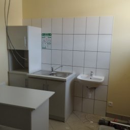 Michał Paszak - Umywalka i zlew ze stali nierdzewnej zamontowane na białych płytkach w pomieszczeniu z żółtymi ścianami, widoczna szafka i skrzynka z narzędziami na podłodze.