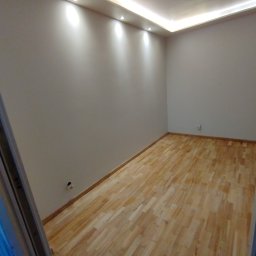 Michał Paszak - Wykończony pokój z parkietem, świeżo pomalowanymi ścianami i oświetleniem sufitowym LED. Widoczny fragment ościeżnicy drzwi. Gniazdko elektryczne na ścianie.