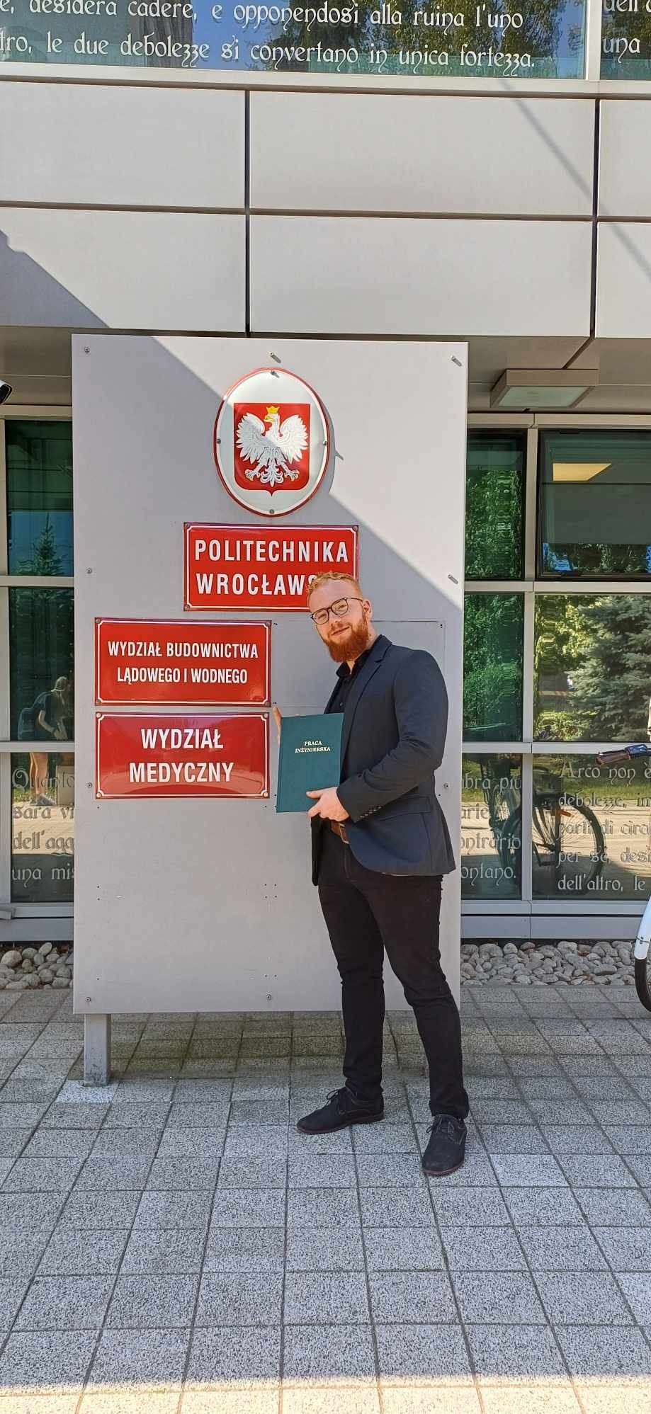 Młody mężczyzna w garniturze, z uśmiechem prezentuje pracę inżynierską na tle budynku Politechniki Wrocławskiej, obok tablic z nazwami wydziałów Budownictwa Lądowego i Wodnego oraz Medycznego.