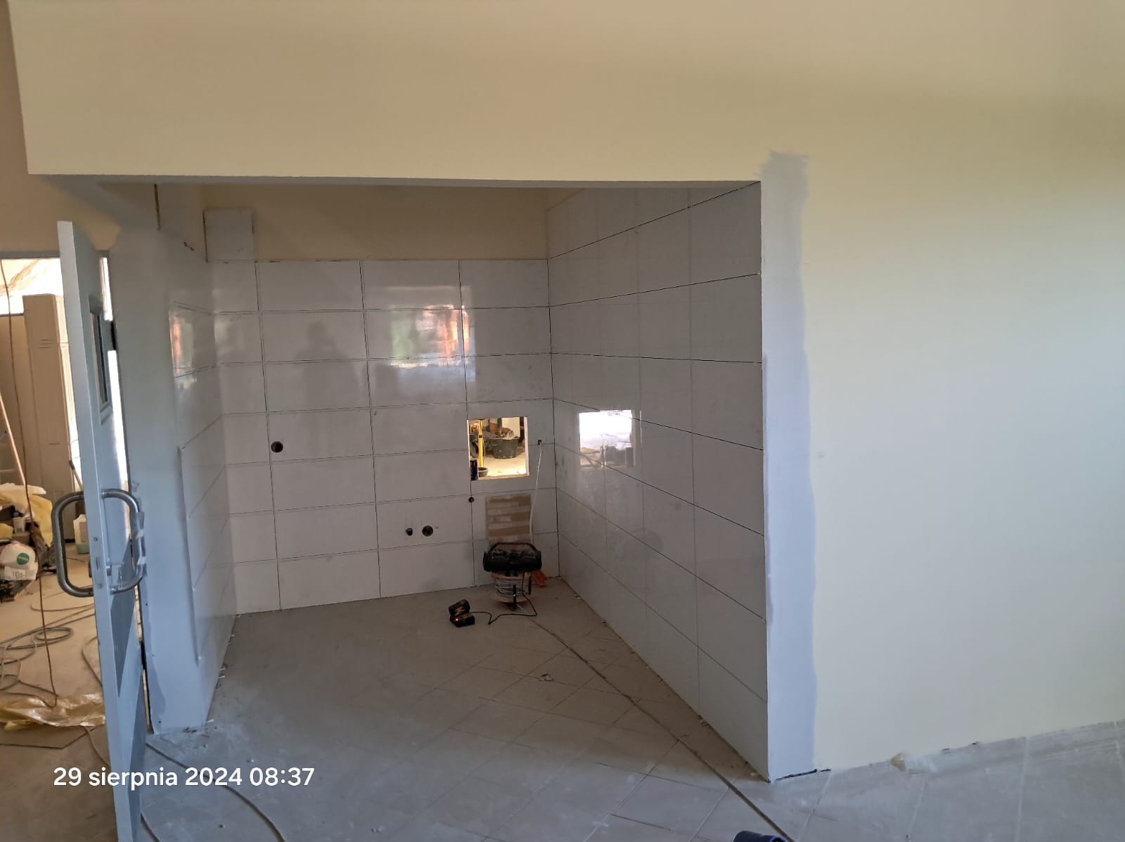 Wnętrze łazienki w trakcie prac wykończeniowych, widoczne białe płytki na ścianach, przygotowane otwory na instalacje, odkurzacz przemysłowy na podłodze, ściany częściowo pomalowane na kremowo...