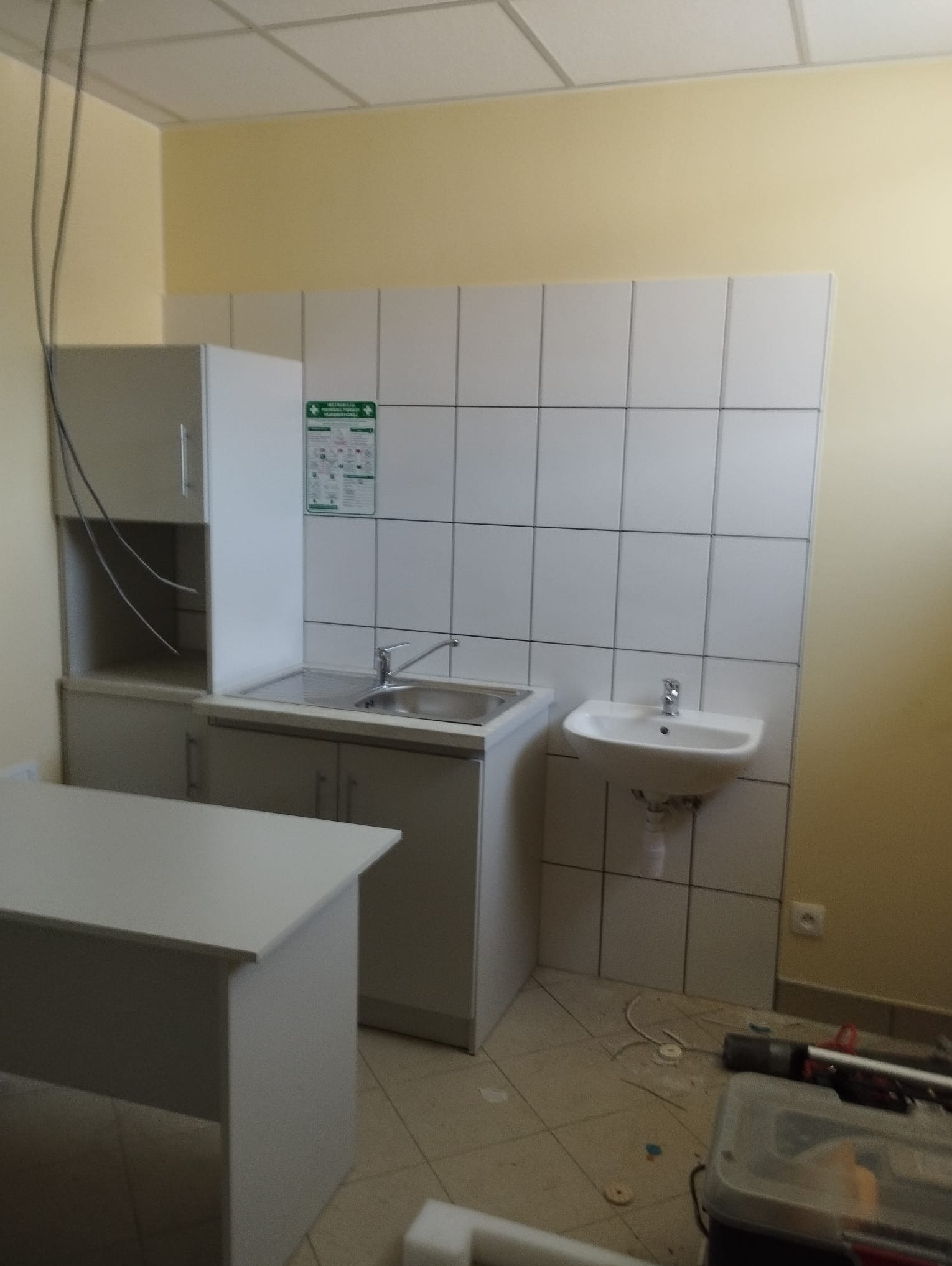 Umywalka i zlew ze stali nierdzewnej zamontowane na białych płytkach w pomieszczeniu z żółtymi ścianami, widoczna szafka i skrzynka z narzędziami na podłodze.