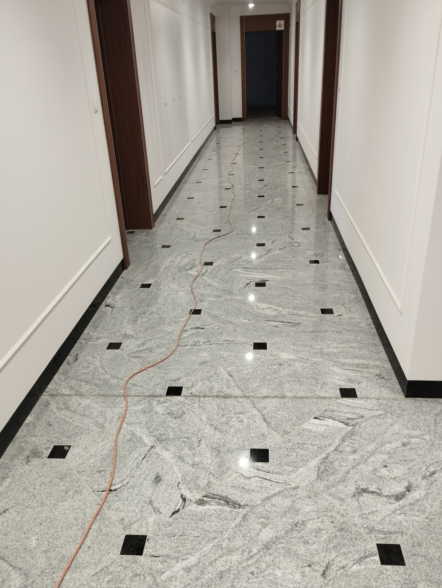 Wykończony korytarz z szarymi płytkami podłogowymi z czarnymi akcentami, widoczny kabel na podłodze.