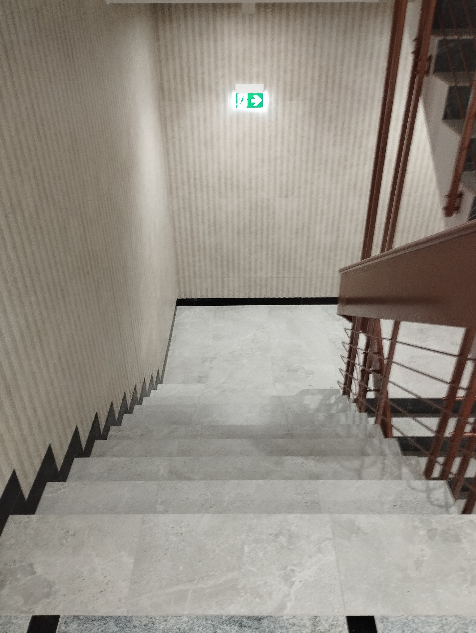 Widok z góry na schody wyłożone jasnoszarymi płytkami z widocznym oznakowaniem drogi ewakuacyjnej na ścianie.