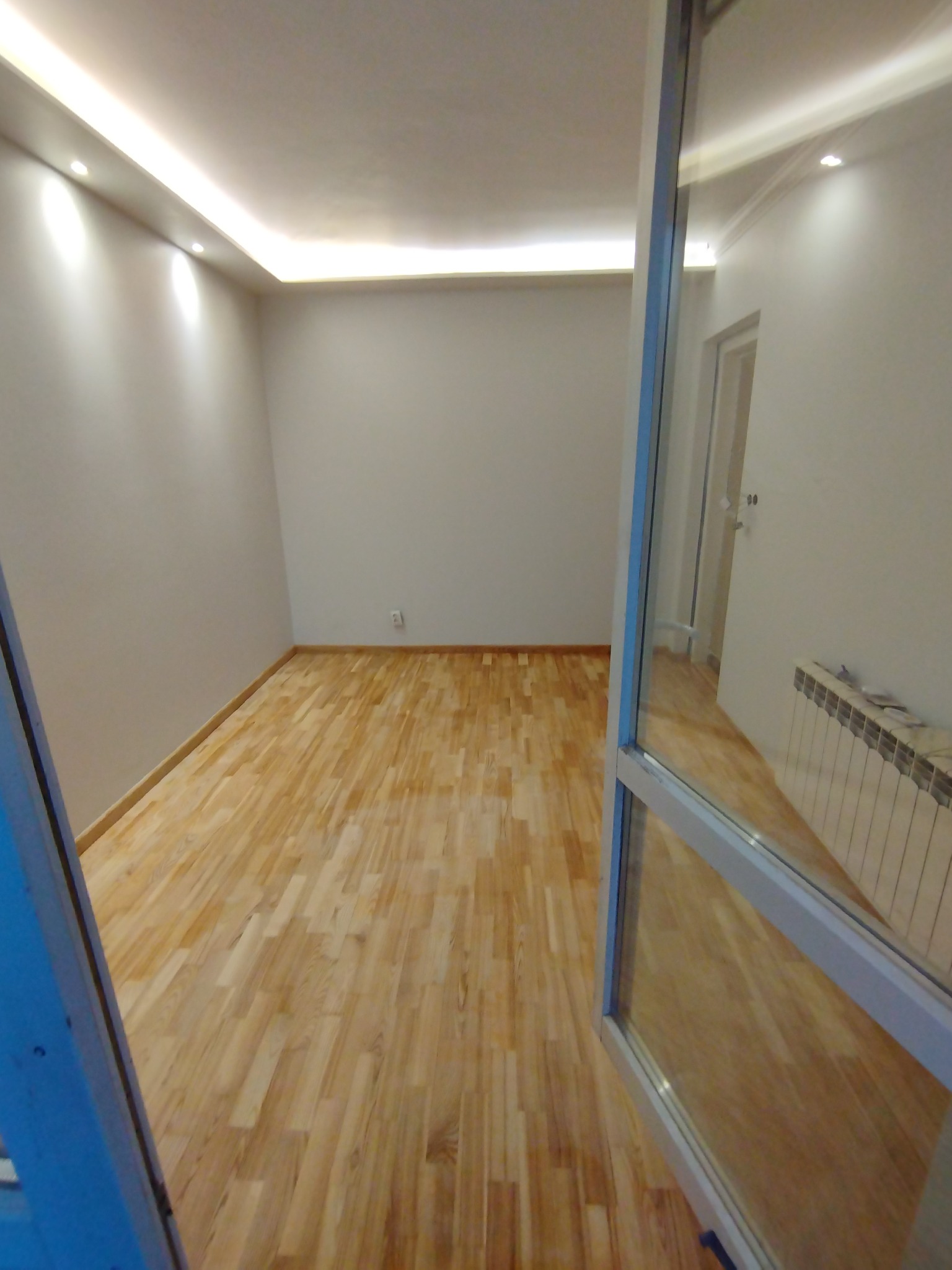 Jasne, puste pomieszczenie z drewnianą podłogą i oświetleniem sufitowym LED, widziane przez otwarte drzwi balkonowe z odbiciem wnętrza.