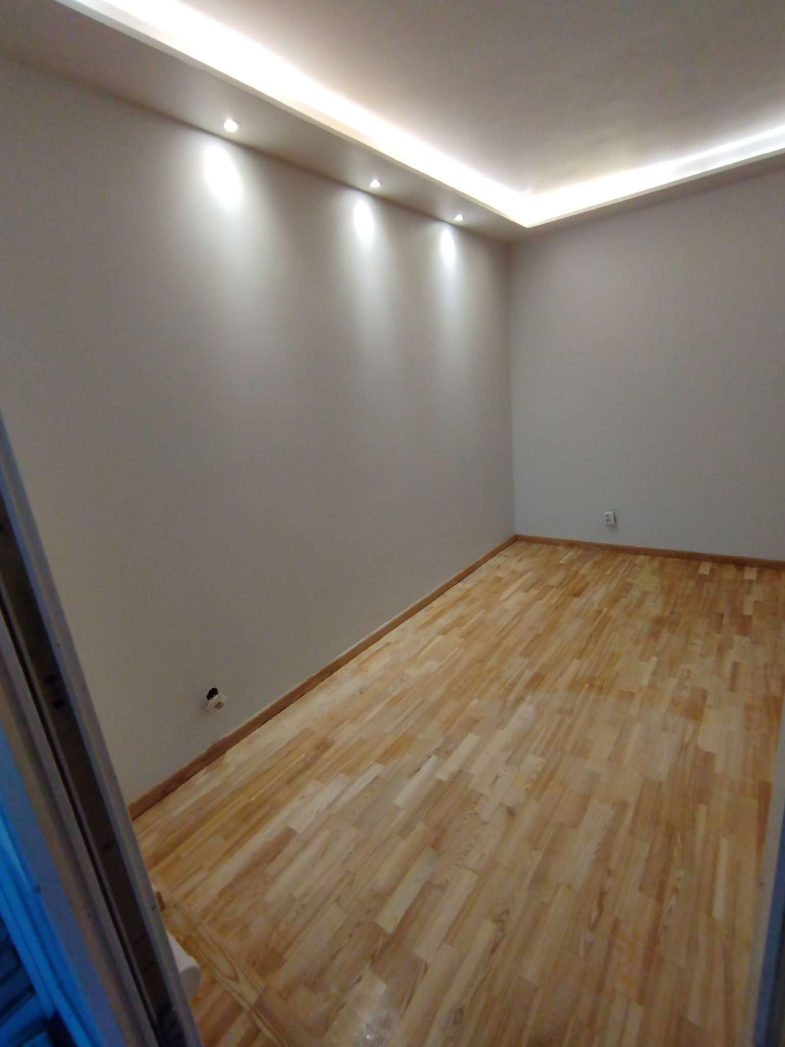 Wykończony pokój z parkietem, świeżo pomalowanymi ścianami i oświetleniem sufitowym LED. Widoczny fragment ościeżnicy drzwi. Gniazdko elektryczne na ścianie.