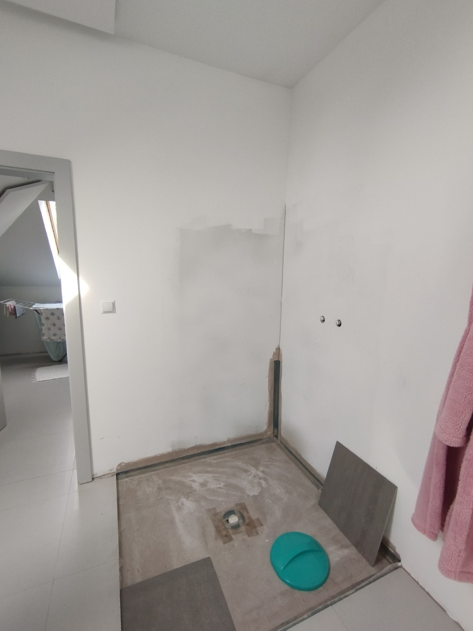 Łazienka w trakcie remontu: przygotowane podłoże pod prysznic, fragmenty płytek, widoczne instalacje. Ściany białe, surowe, z śladami po pracach wykończeniowych.