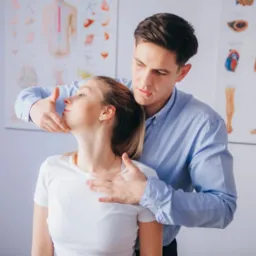 Mężczyzna w niebieskiej koszuli delikatnie manipuluje szyją pacjentki w białym T-shircie, z plakatami anatomicznymi w tle.