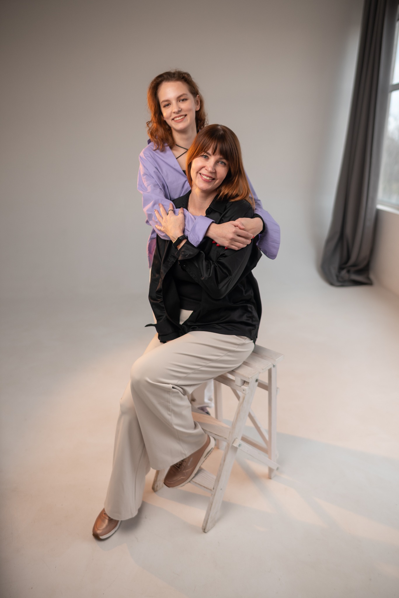 Dwie uśmiechnięte kobiety w studyjnym oświetleniu: jedna siedzi na białym stołku w czarnej koszuli i beżowych spodniach, druga stoi za nią w liliowej koszuli, obejmując ją ramionami. W tle szara...