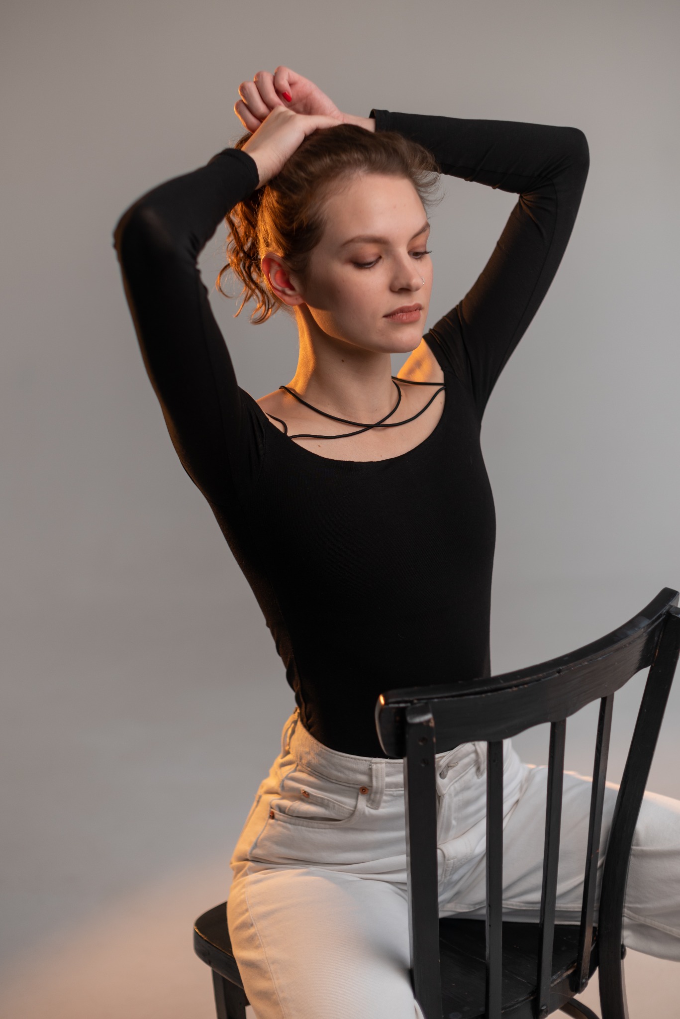 Portret kobiety siedzącej na czarnym krześle, w czarnym body i białych jeansach, z rękami uniesionymi do góry układającej włosy, w studiu oświetlonym ciepłym światłem.