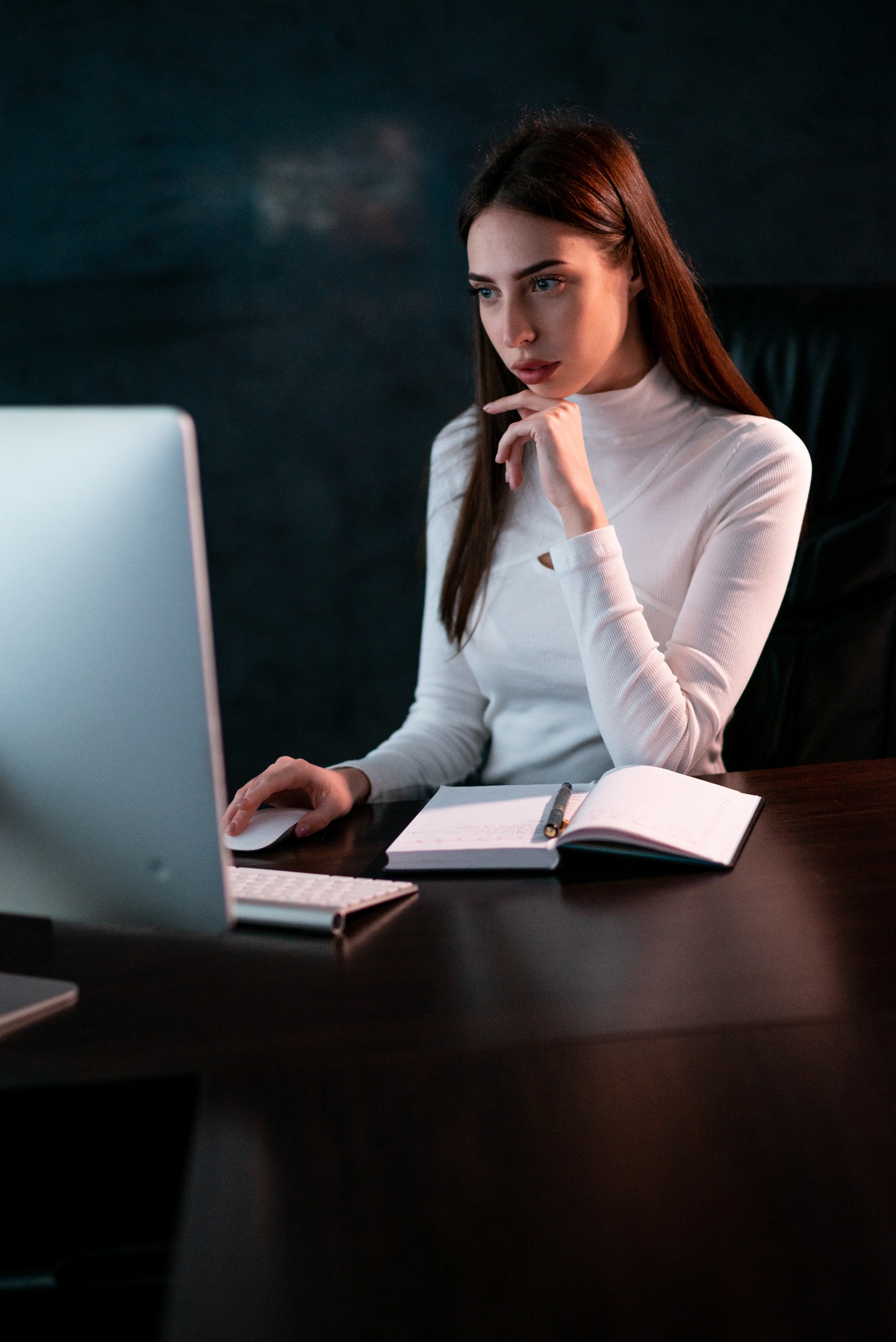 Kobieta w białym golfie siedzi przy biurku, skupiona na ekranie komputera, z dłonią na myszce i otwartym notatniku obok.