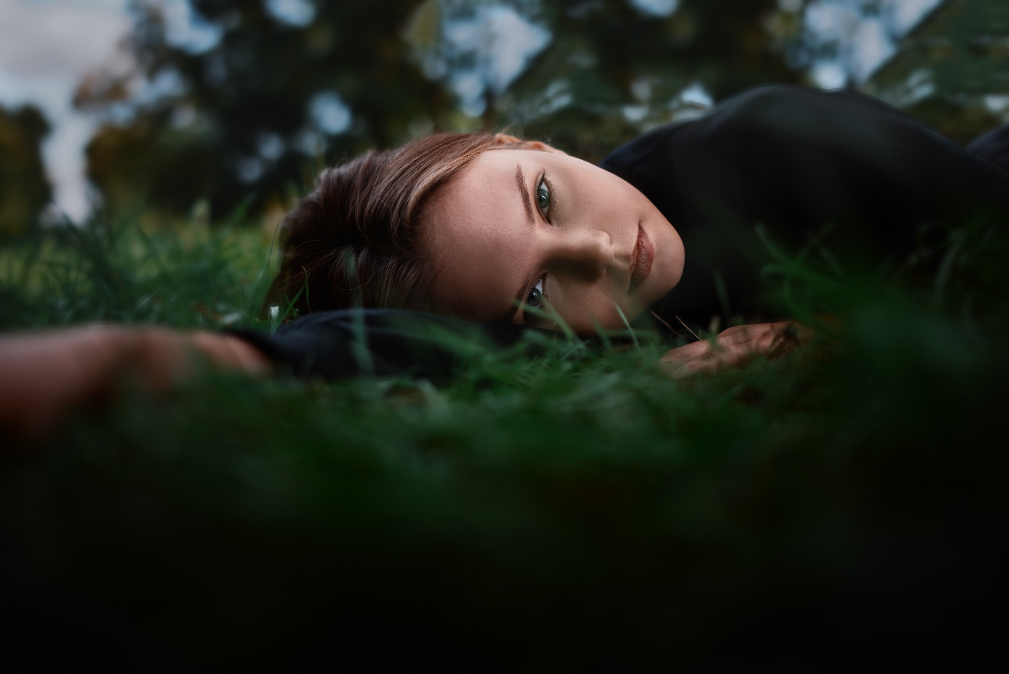 Portret kobiety z zielonymi oczami leżącej na trawie w ciemnym ubraniu, z rozproszonym tłem drzew.