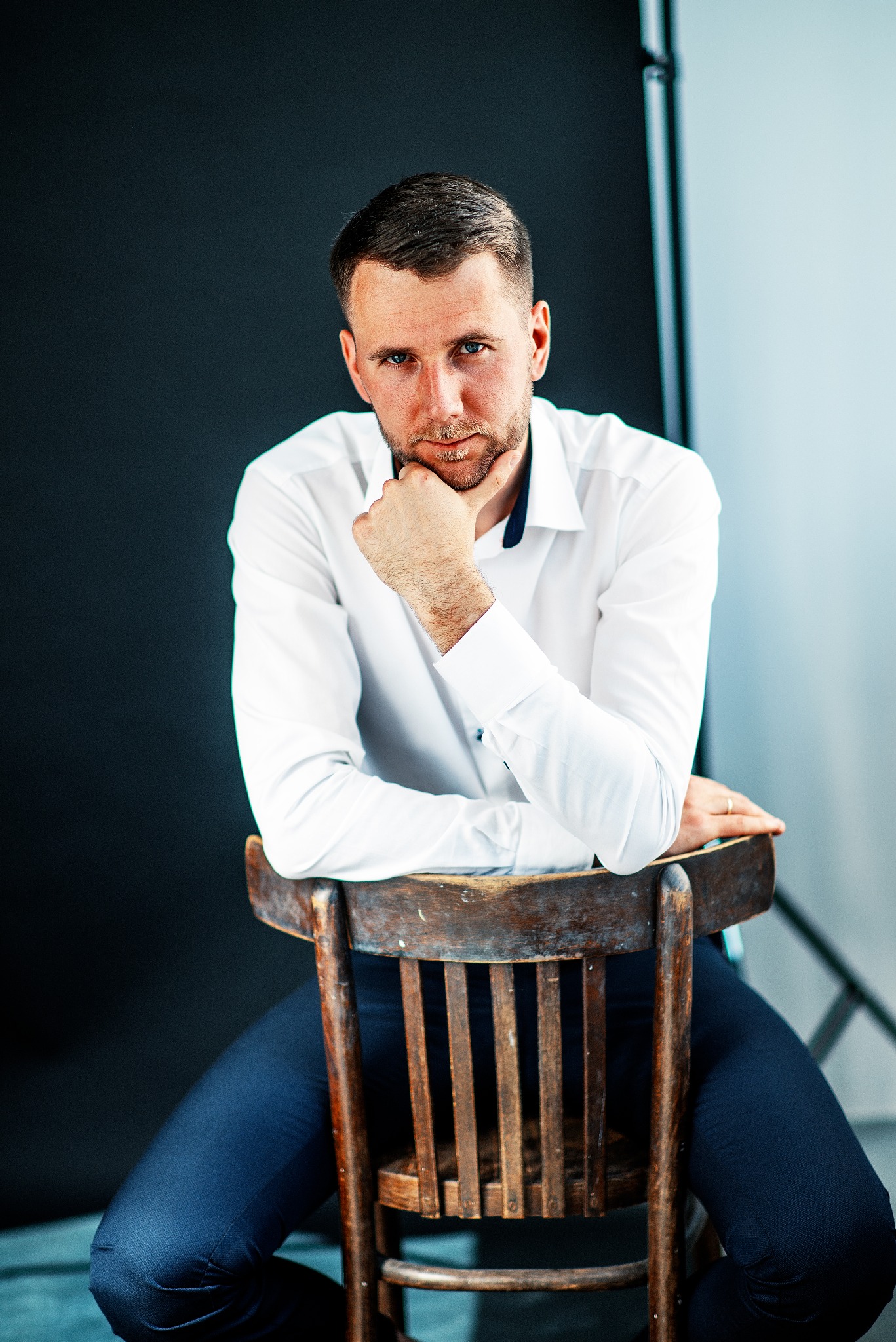Mężczyzna w białej koszuli i granatowych spodniach siedzi na drewnianym krześle, opierając brodę na dłoni, na tle czarno-białego studia.