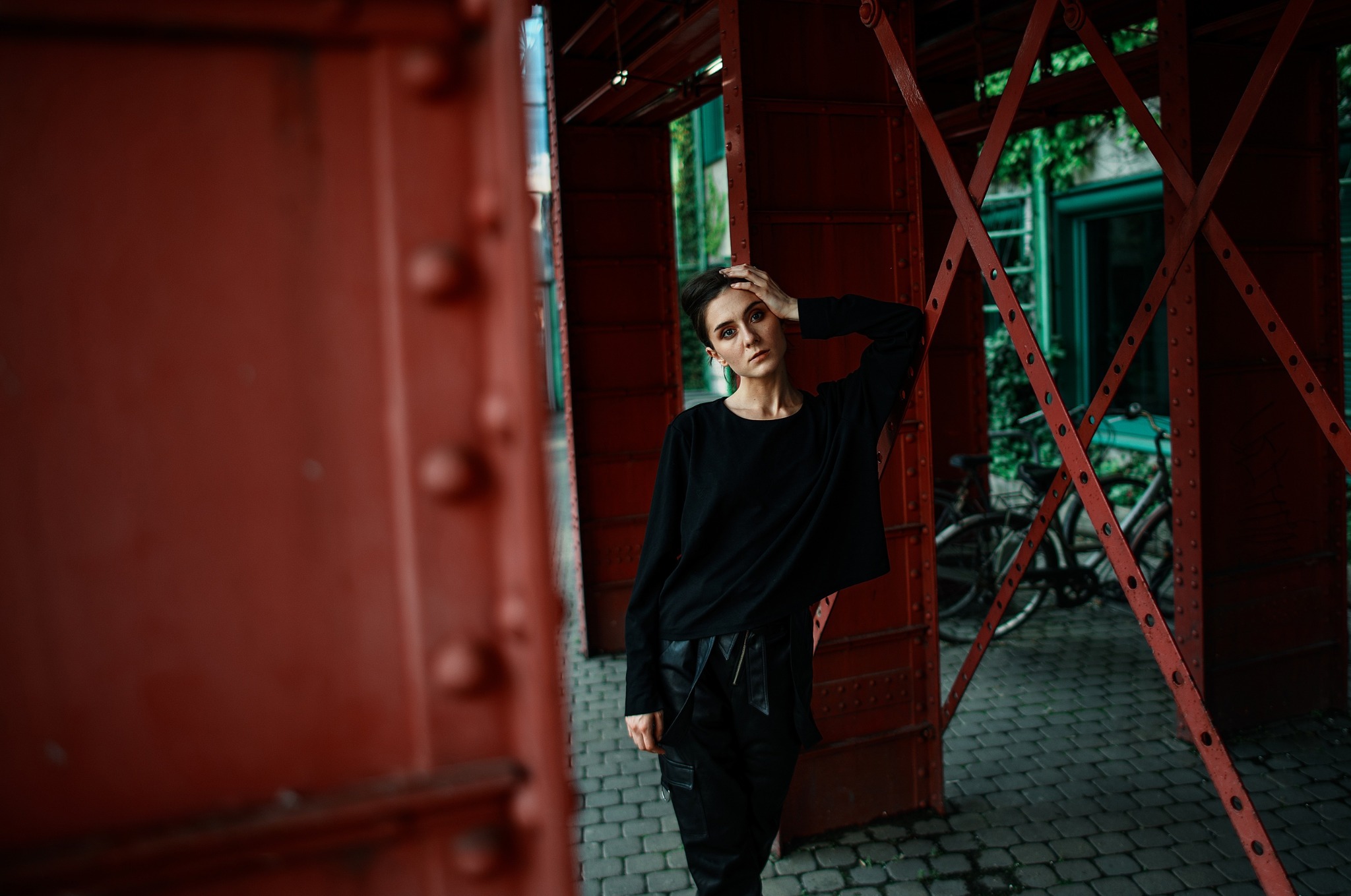 Portret kobiety w czarnym ubraniu na tle czerwonej konstrukcji w Warszawie, z rowerami i zielenią w tle.