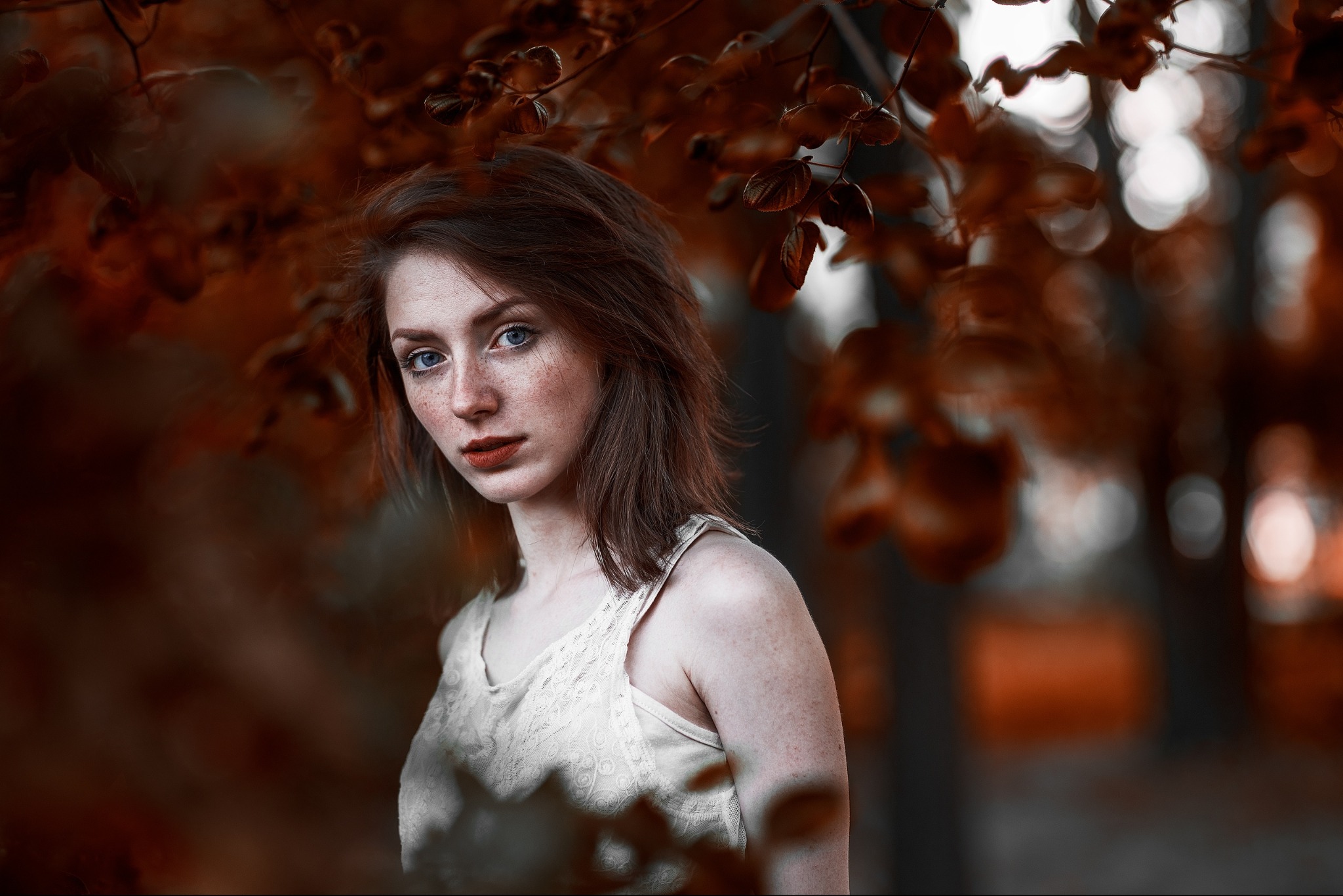 Portret młodej kobiety z piegami i niebieskimi oczami, otoczonej jesiennymi liśćmi w ciepłej tonacji, ubrana w białą koronkową koszulkę na ramiączkach.