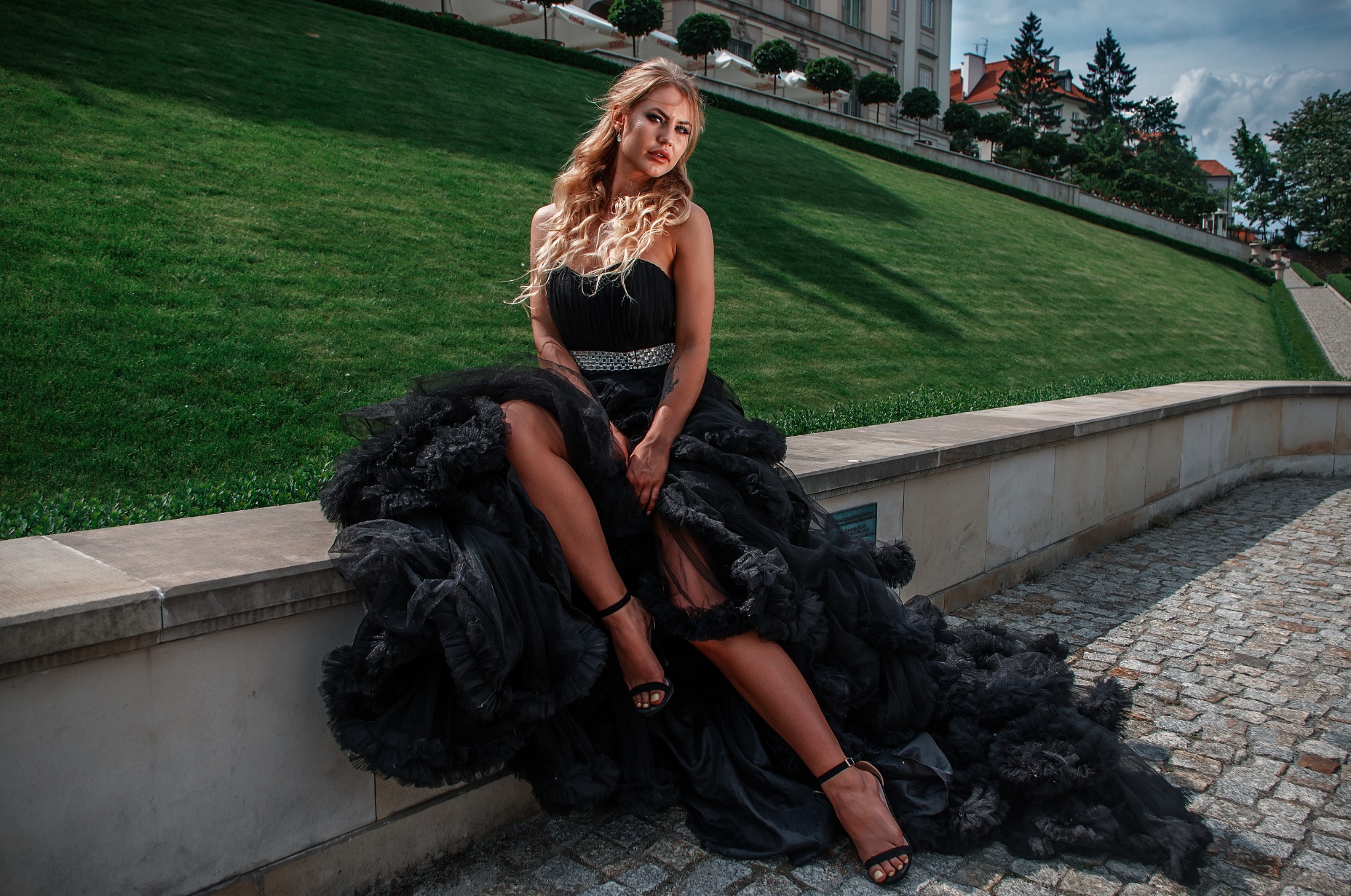Kobieta w czarnej tiulowej sukni z cekinowym pasem, siedząca na kamiennym murku na tle zielonego trawnika i klasycystycznego budynku z ozdobnymi drzewami.