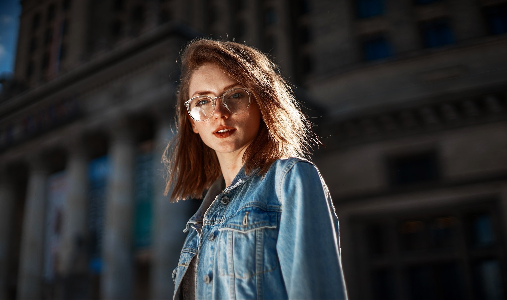 Portret młodej kobiety w okularach, w dżinsowej kurtce na tle architektury miejskiej z kolumnami, oświetlonej popołudniowym słońcem.