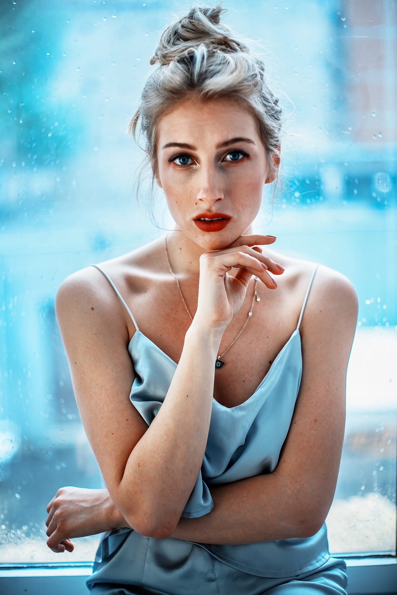 Portret kobiety z włosami upiętymi w kok, w satynowej koszuli na ramiączkach, stojącej na tle okna z kroplami deszczu, z ręką pod brodą i melancholijnym wyrazem twarzy.