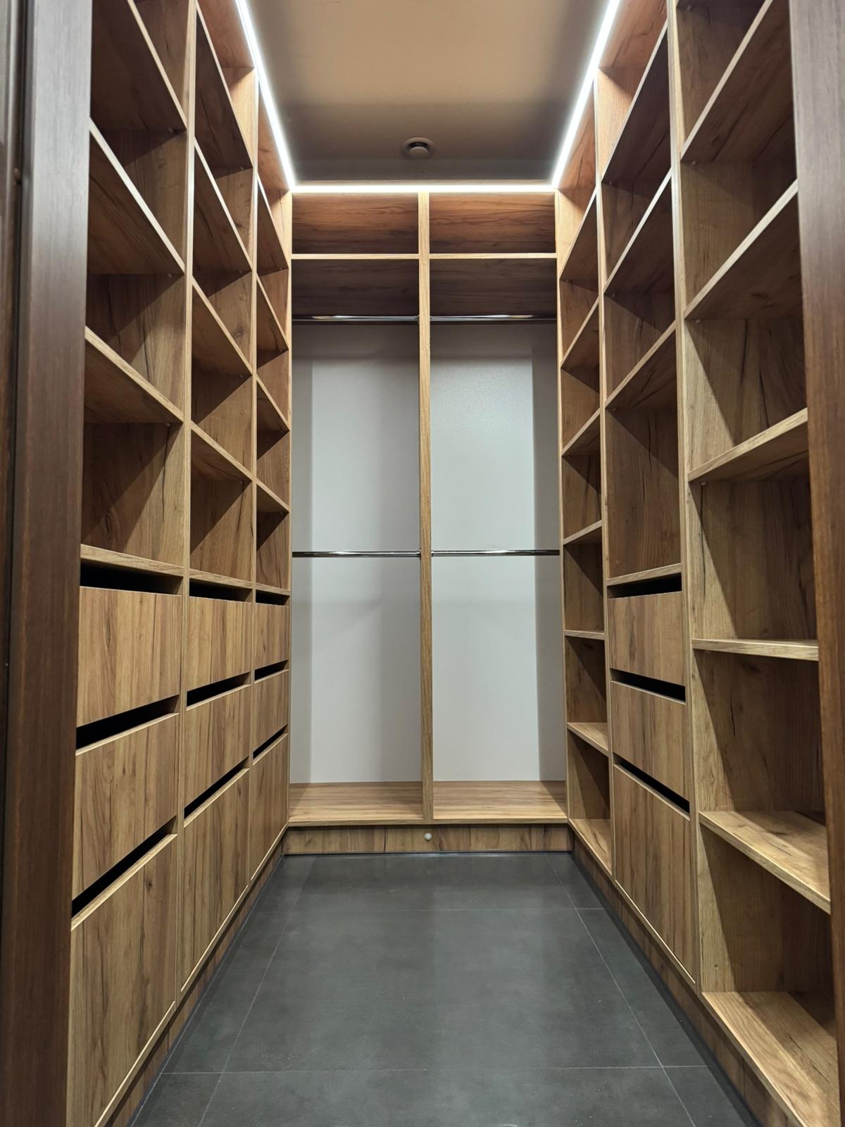 Wnętrze garderoby na wymiar z oświetleniem LED, drewnopodobne fronty szafek z minimalistycznymi uchwytami, szare płytki podłogowe.