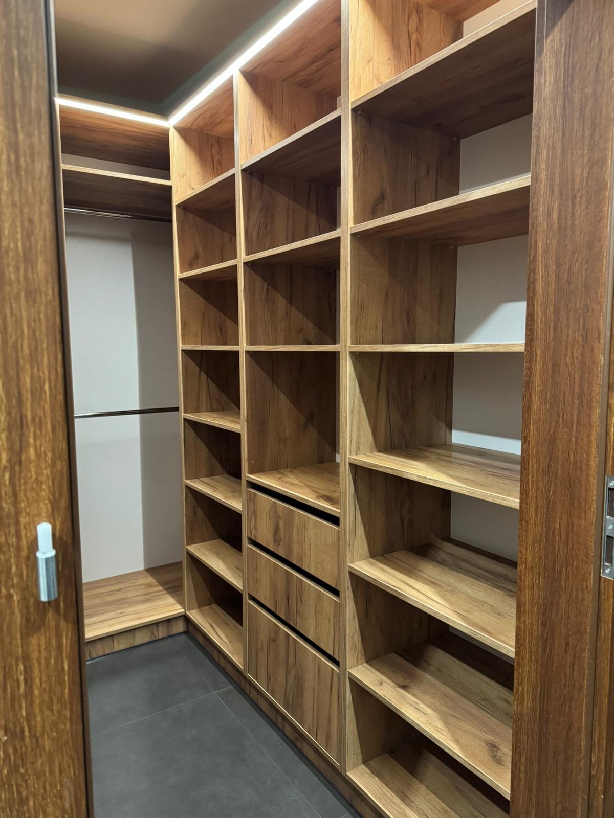 Wnętrze garderoby z drewnopodobnymi półkami i szufladami, oświetlenie LED w górnej części, widok z otwartych drzwi.