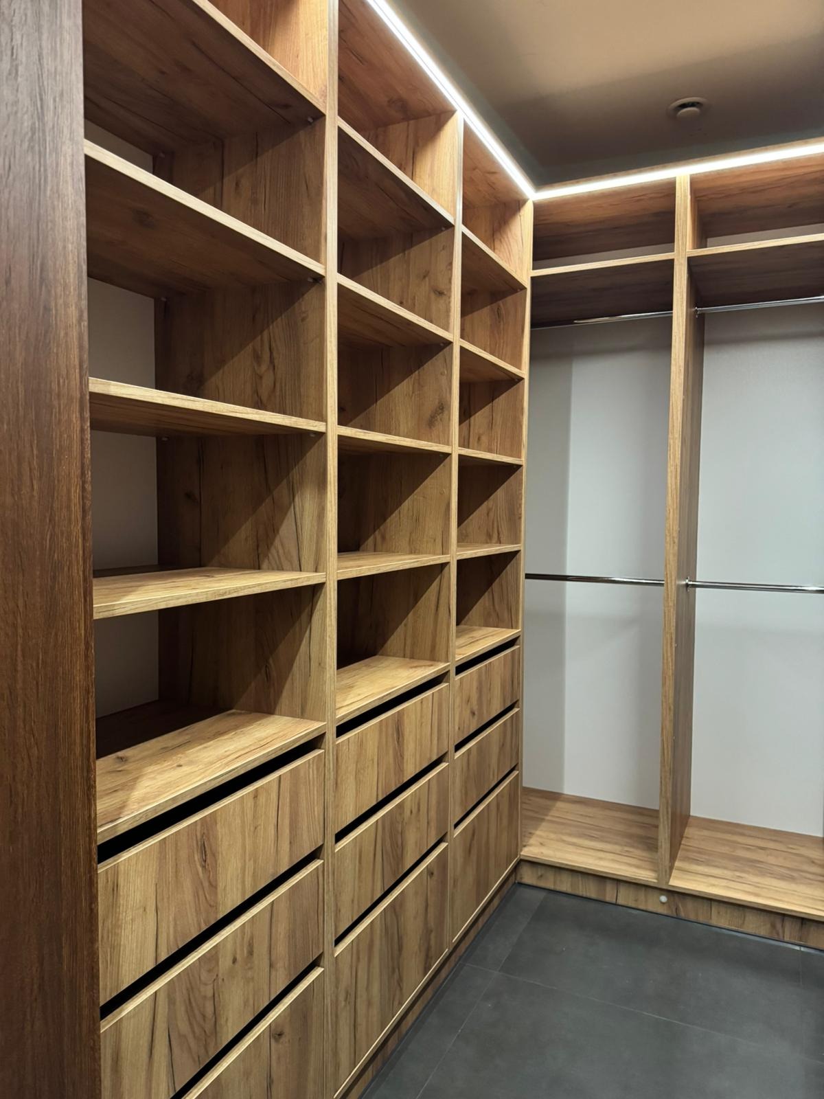 Wnętrze garderoby z drewnianymi półkami i szufladami, oświetlenie LED w górnej części, widok z perspektywy podłogi z szarymi płytkami.