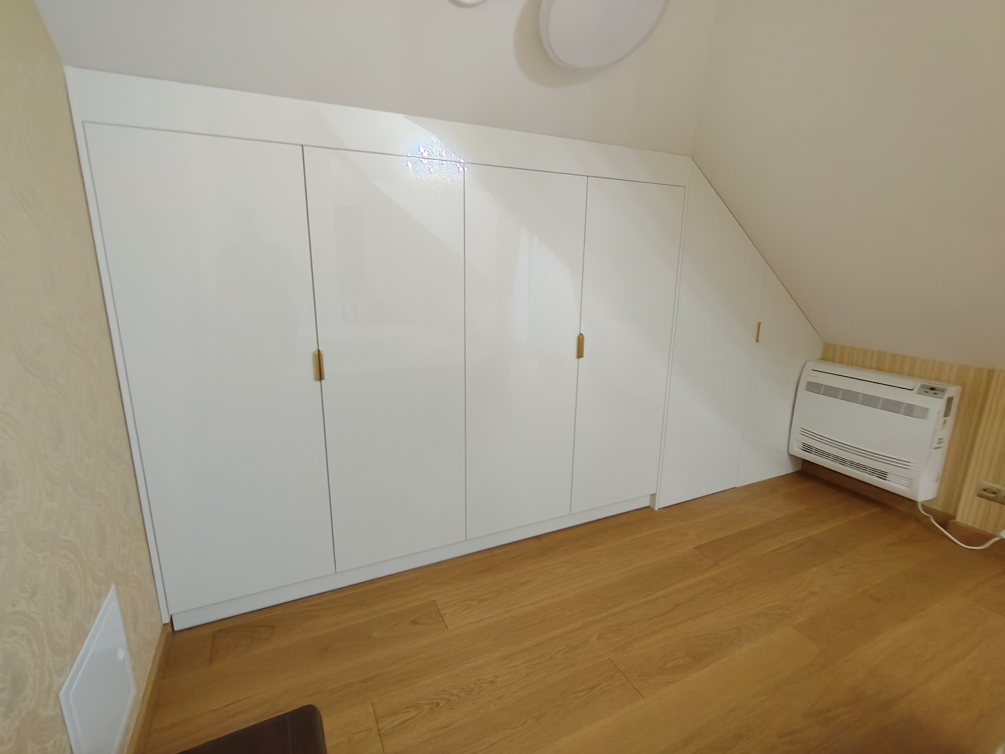 Biała, lakierowana szafa wnękowa pod skosem dachu, z czterema drzwiami i złotymi uchwytami, na drewnianej podłodze, z grzejnikiem obok.