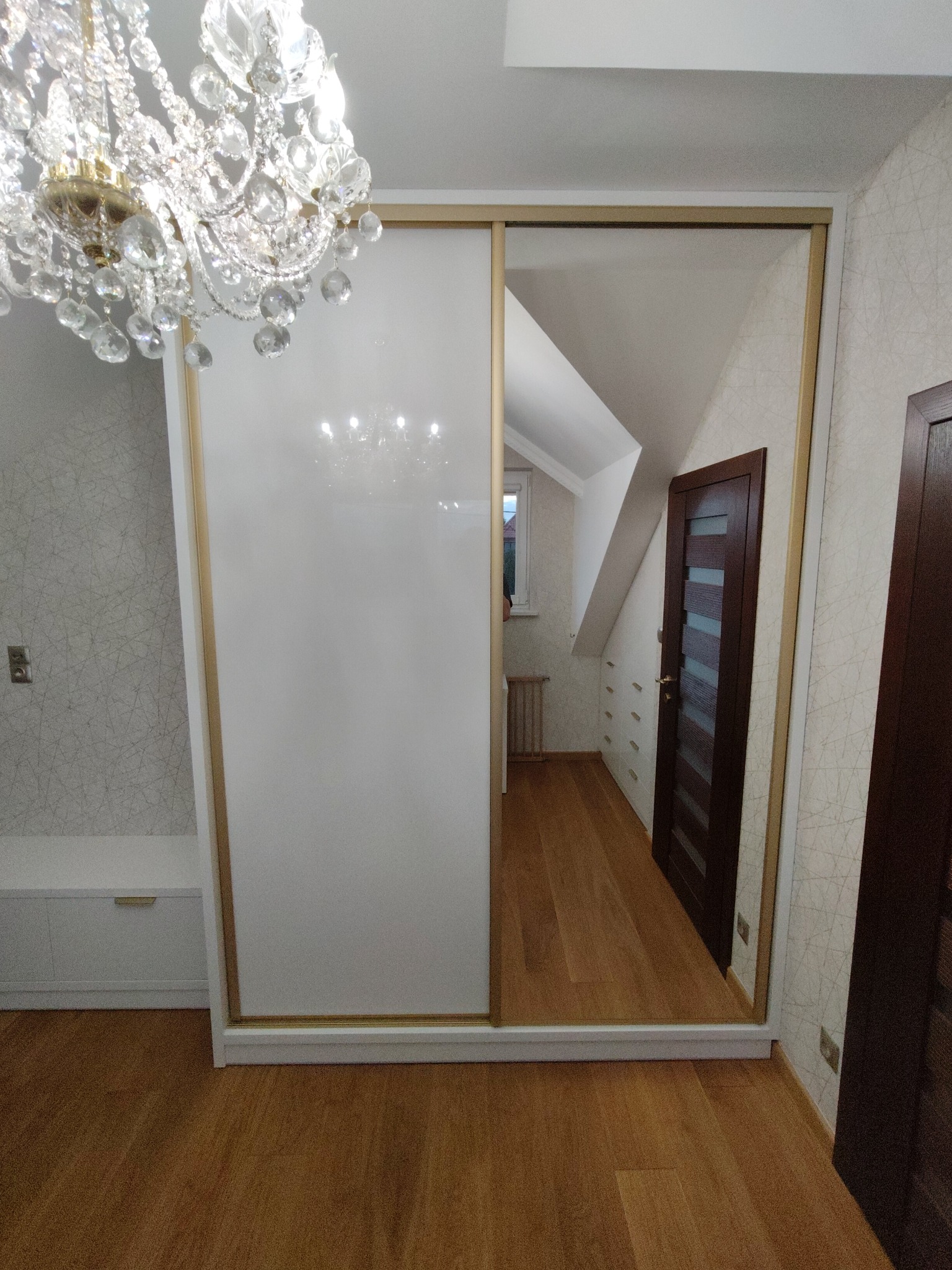 Szafa wnękowa z przesuwnymi drzwiami: biały panel i lustro, widoczne odbicie pokoju na poddaszu, oświetlenie kryształowym żyrandolem.