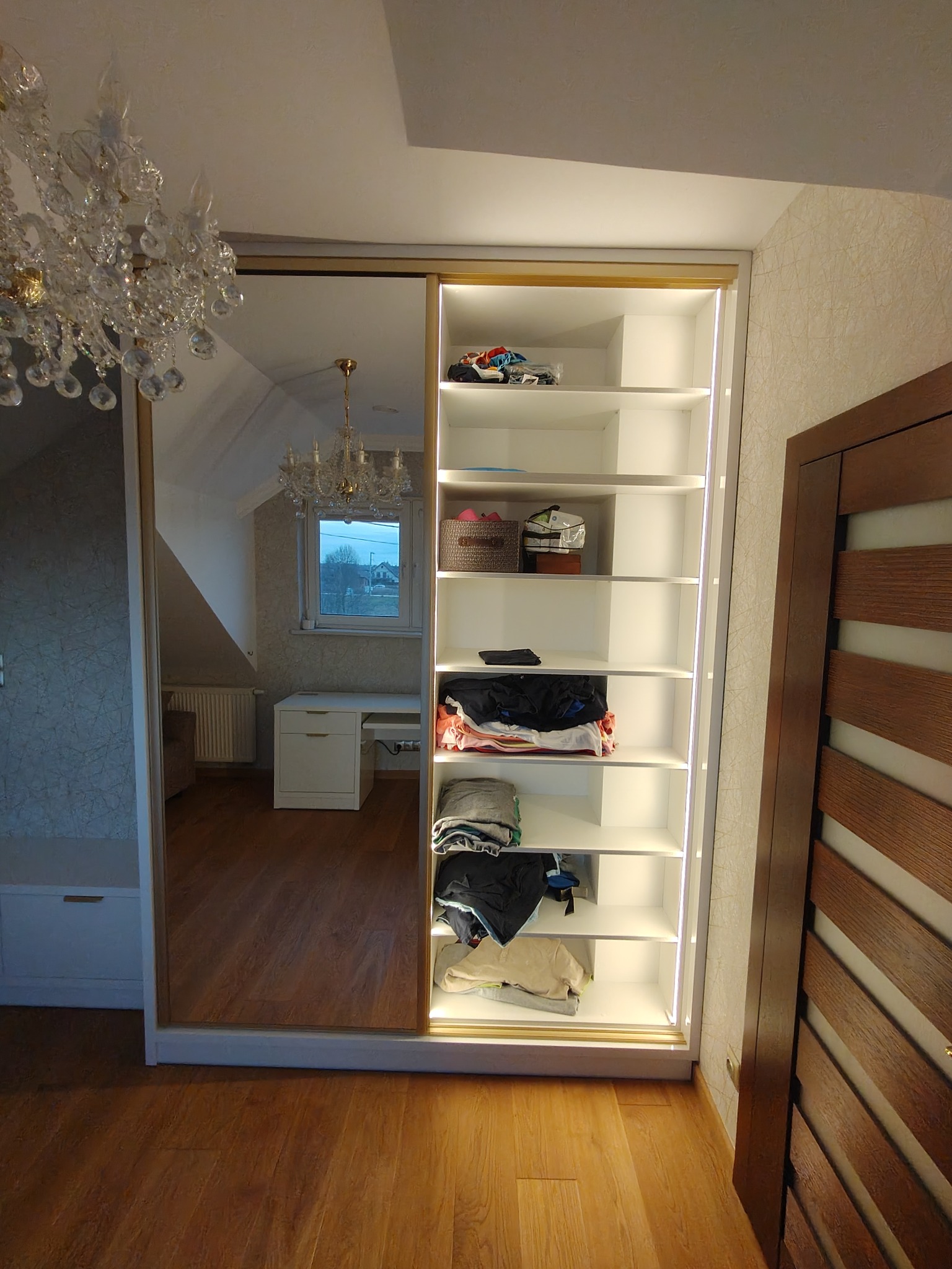 Biała szafa wnękowa z przesuwnymi drzwiami lustrzanymi, oświetleniem LED i półkami wypełnionymi ubraniami, widoczna w odbiciu kryształowy żyrandol.