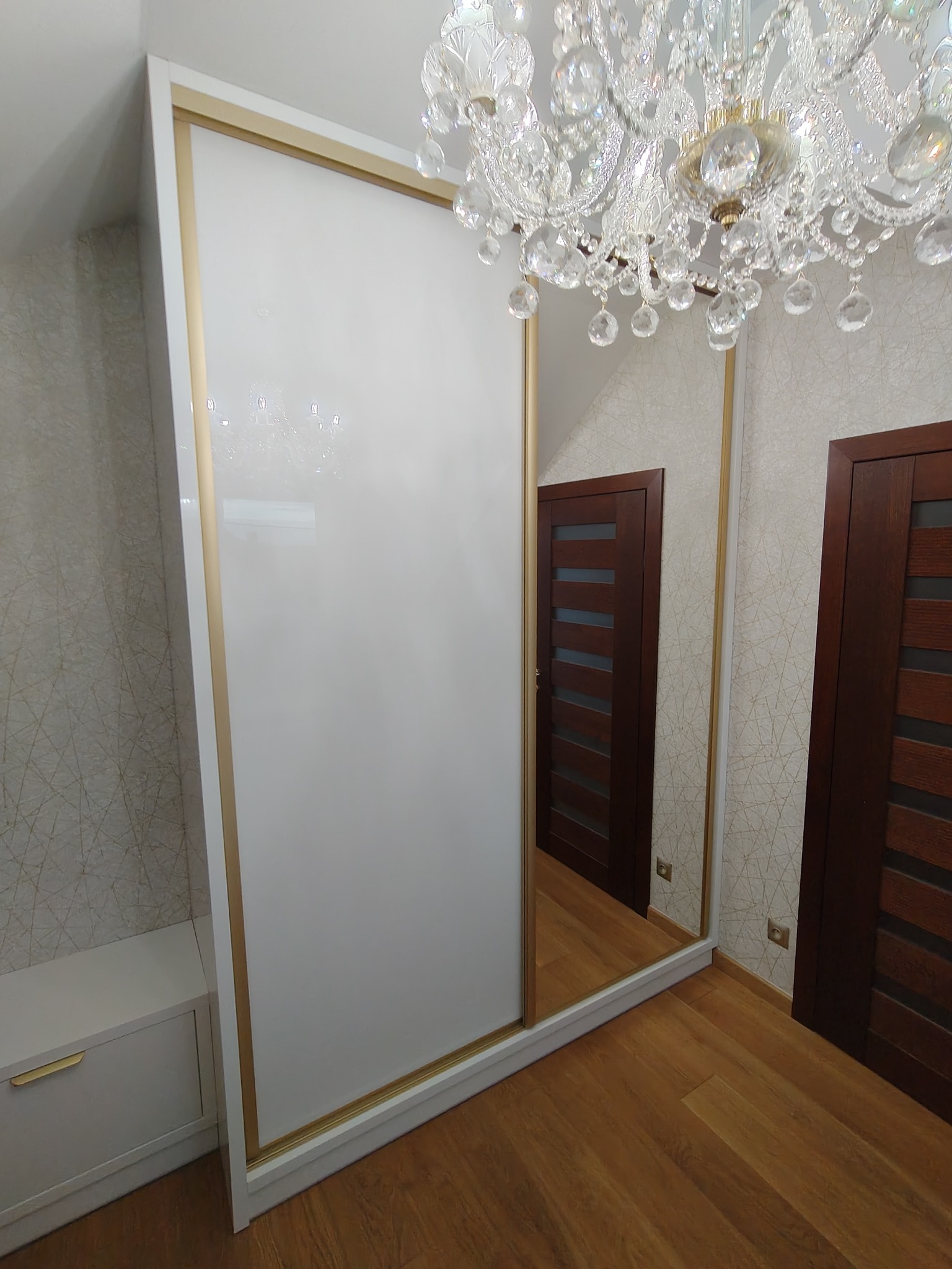Biała szafa przesuwna z lustrzanymi drzwiami i złotymi akcentami, uzupełniona kryształowym żyrandolem, odbijająca drzwi w lustrze.