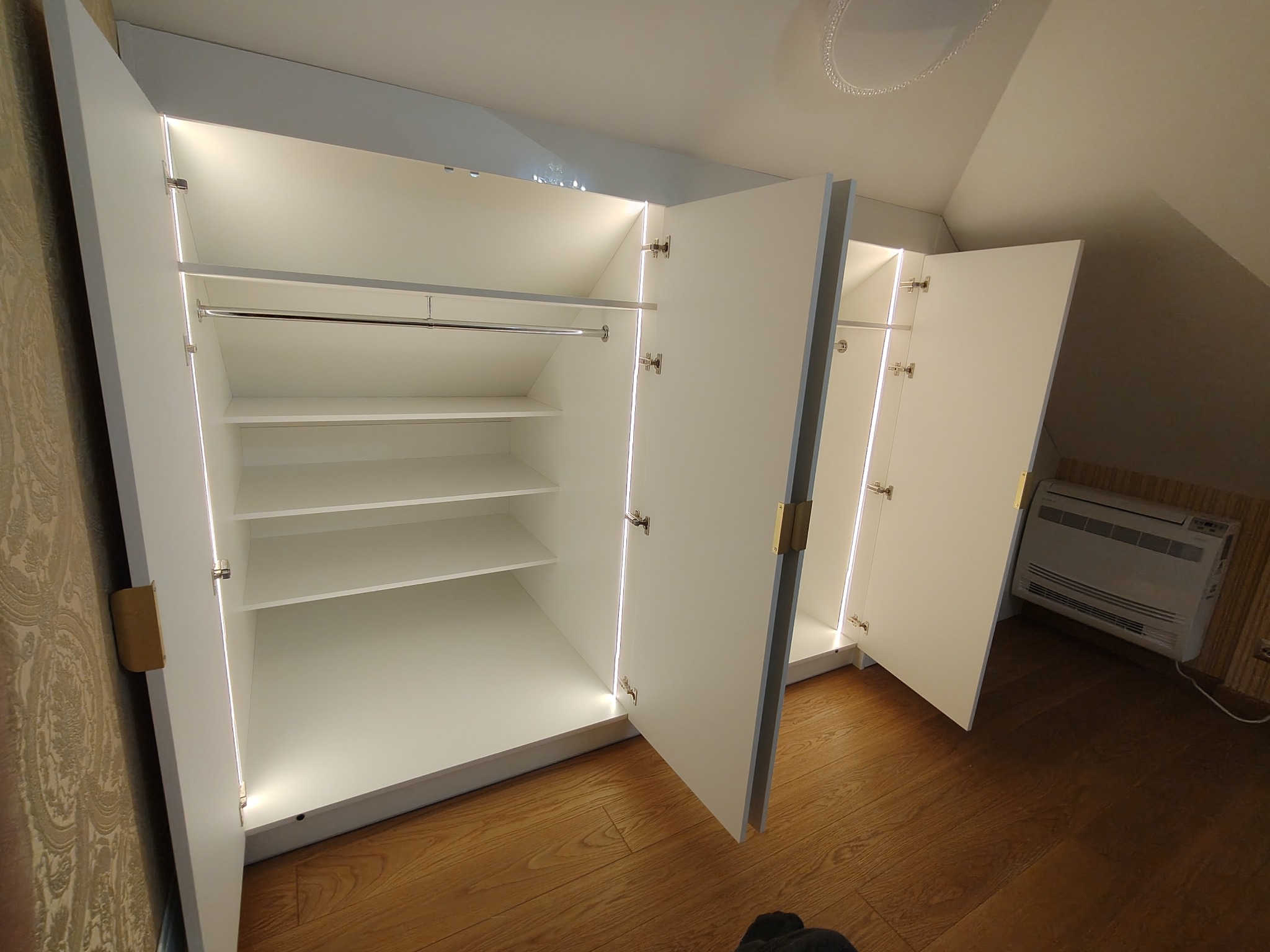 Biała szafa wnękowa z oświetleniem LED, otwarte drzwi ukazują puste półki i drążek na ubrania, widoczna podłoga z paneli i fragment ściany z wzorzystą tapetą.