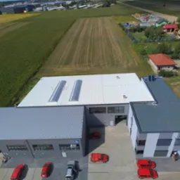 Zdjęcie z drona przedstawiające nowoczesną halę produkcyjną z szarej blachy trapezowej z oknami i bramami, z białym dachem z świetlikami dachowymi, otoczoną polami i zabudową jednorodzinną,...