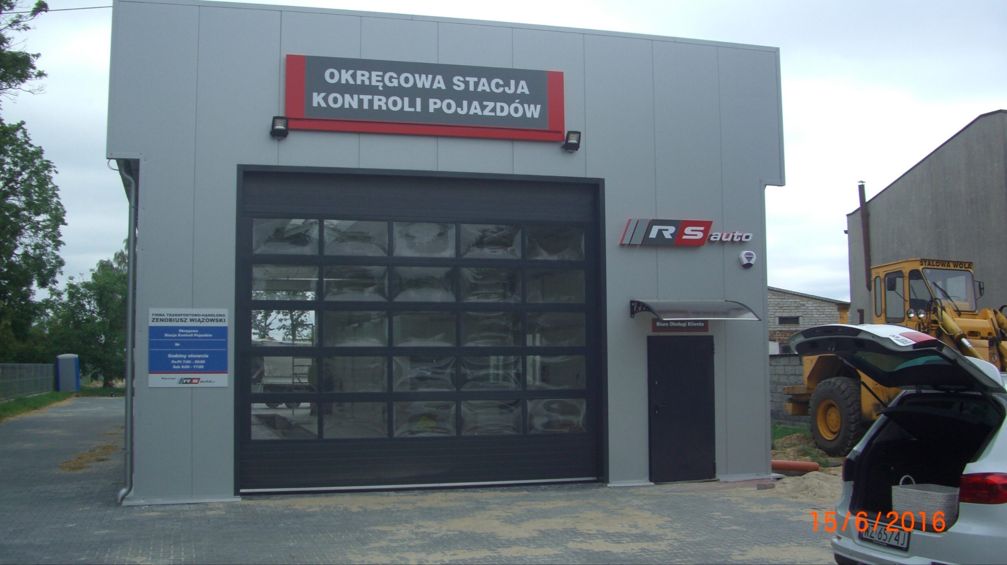 Budynek Okręgowej Stacji Kontroli Pojazdów z szarą elewacją, bramą segmentową z przeszkleniami, logo RS auto, widoczny fragment zaparkowanego samochodu i żółtej koparki na zewnątrz.