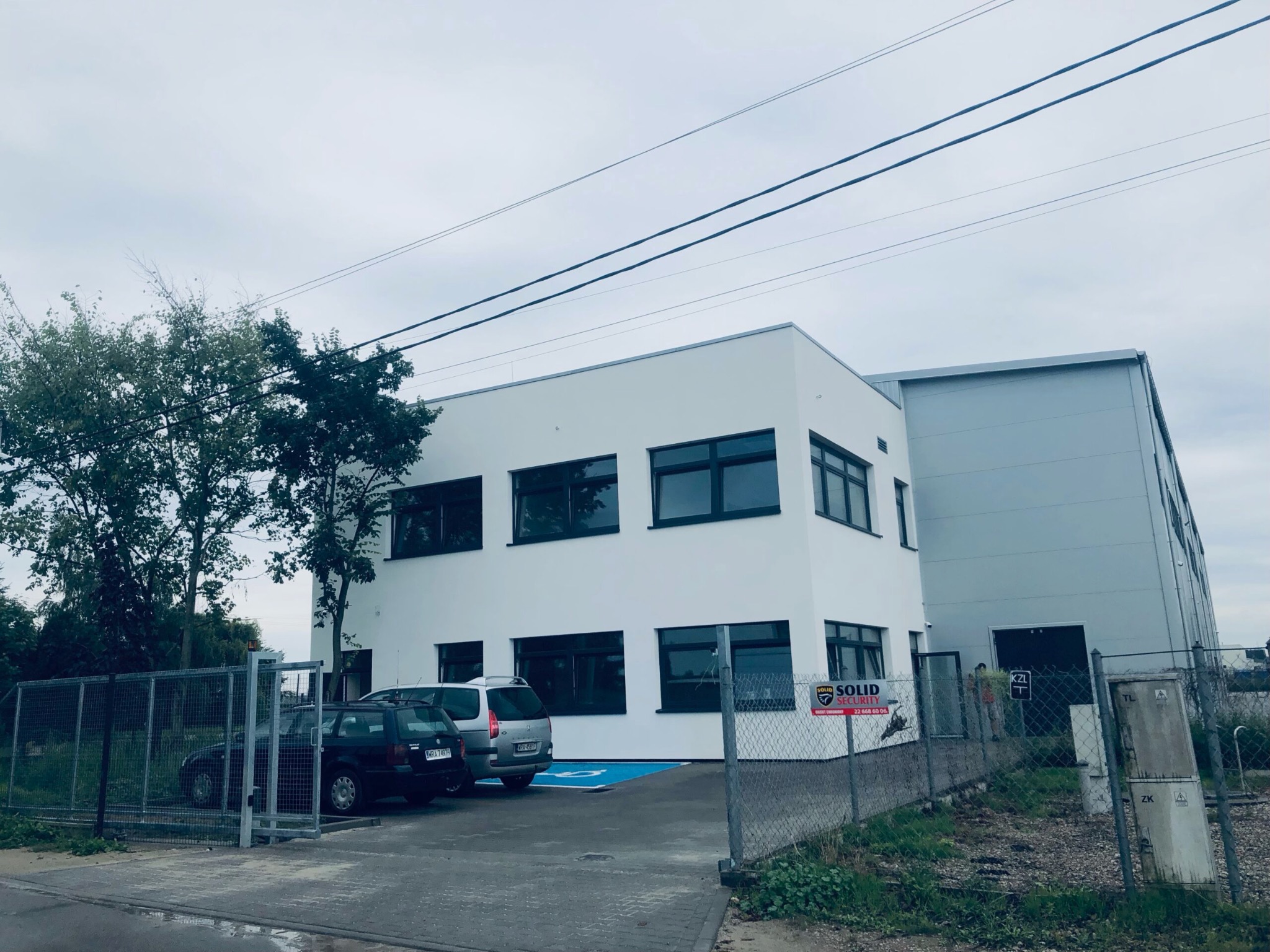 Nowoczesny budynek biurowo-magazynowy z białymi ścianami i ciemnymi oknami, widoczny zza metalowego ogrodzenia, z zaparkowanymi samochodami i niebieskim miejscem parkingowym dla niepełnosprawnych.
