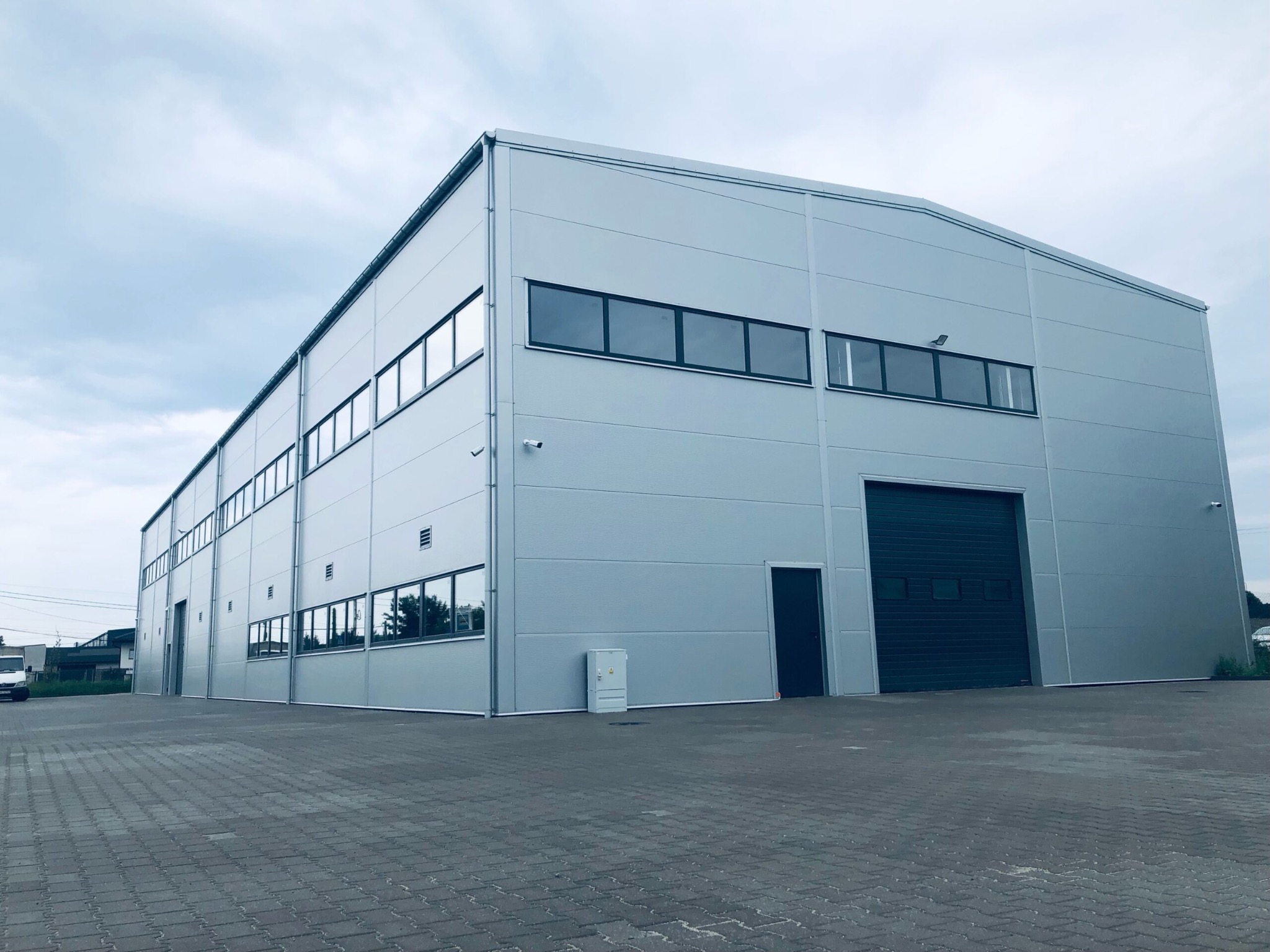 Gotowa hala przemysłowa z szarej blachy trapezowej, z bramą segmentową i oknami, widok z zewnątrz na tle pochmurnego nieba.