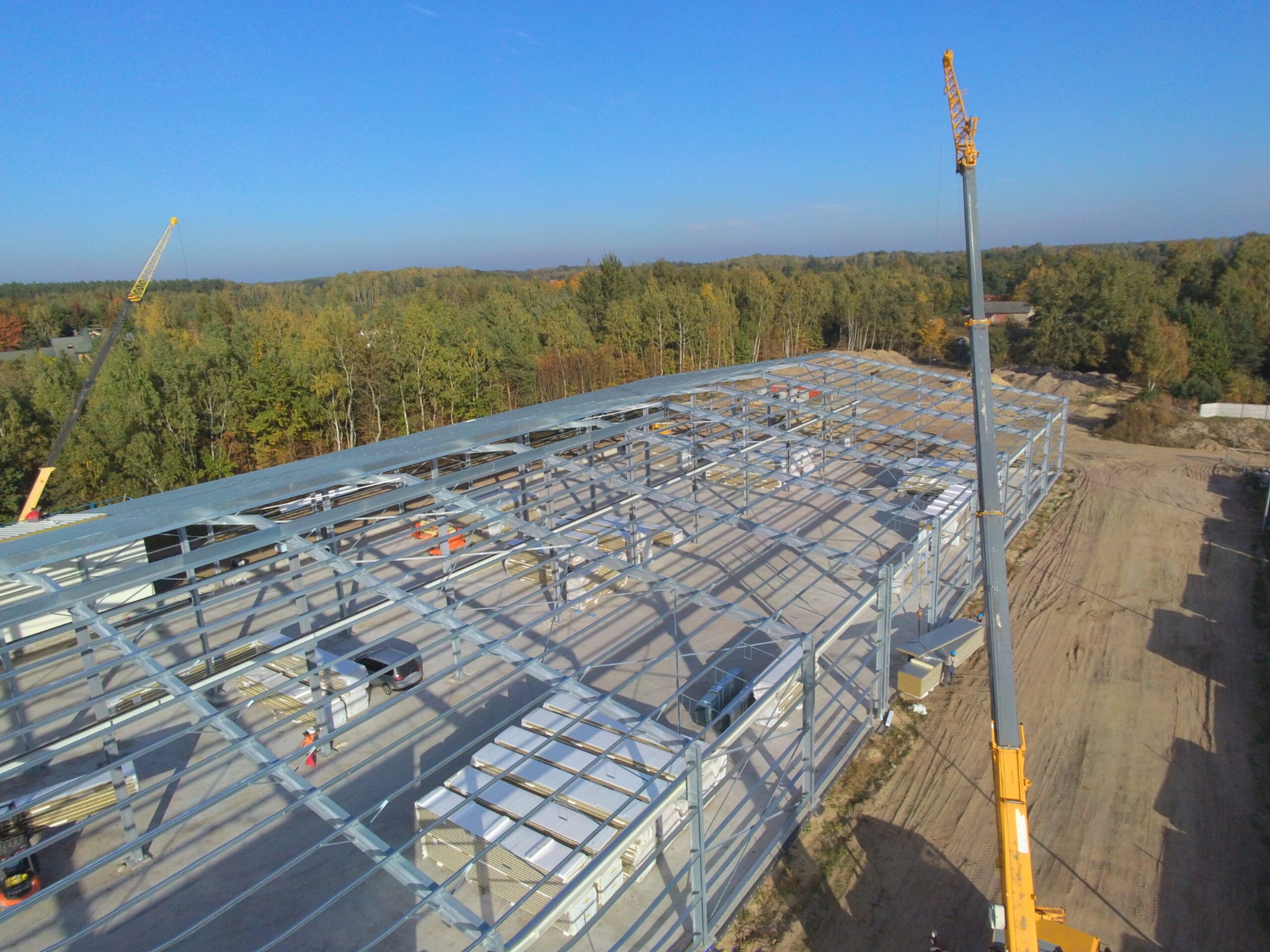 Widok z góry na stalową konstrukcję hali w trakcie budowy, z dwoma dźwigami na placu budowy i stosem płyt warstwowych wewnątrz szkieletu.