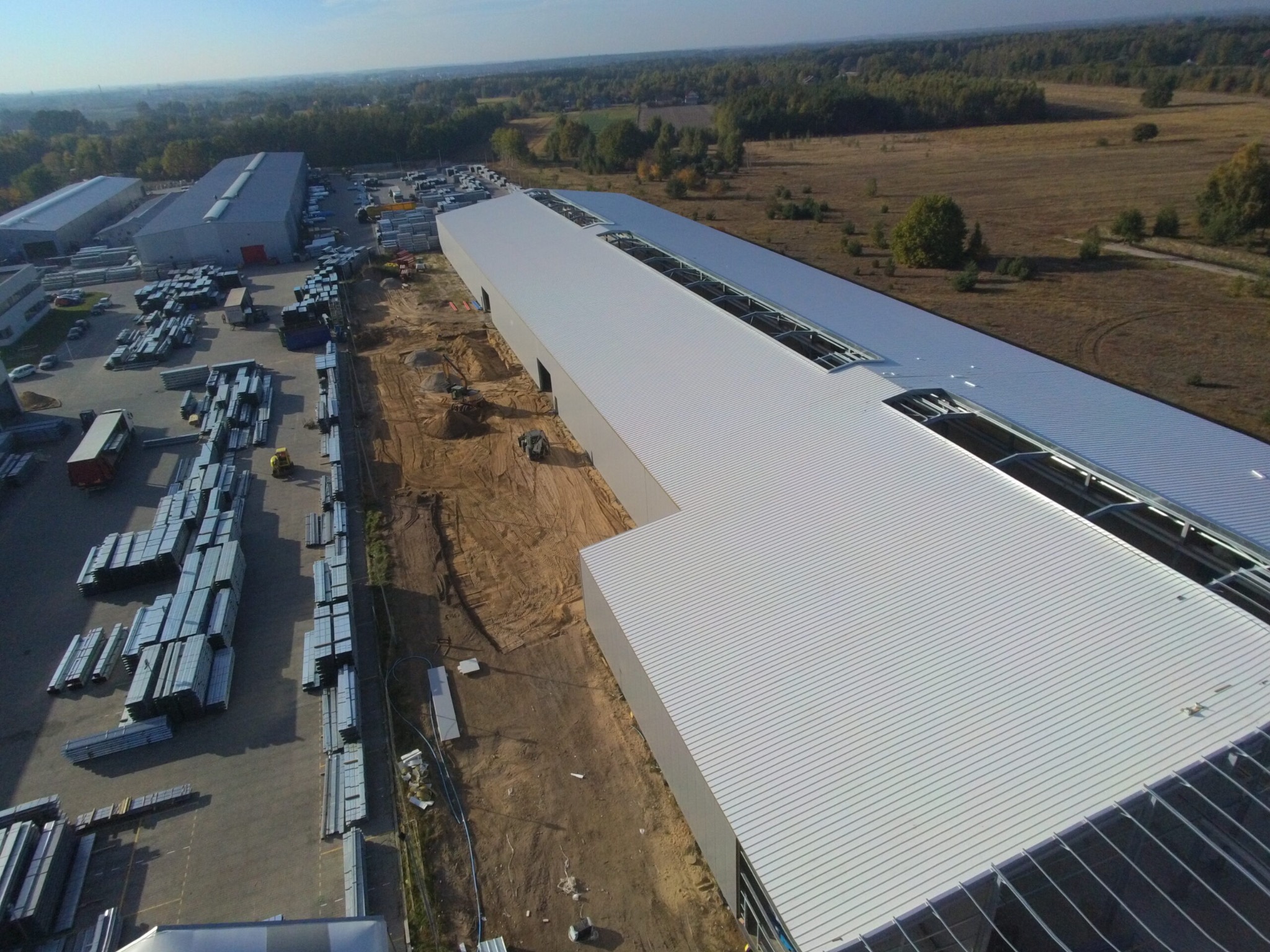Zdjęcie z lotu ptaka przedstawiające budowę hali z jasnoszarą blachą trapezową, widoczne stosy elementów konstrukcyjnych na placu, ziemny teren budowy i okoliczne lasy.