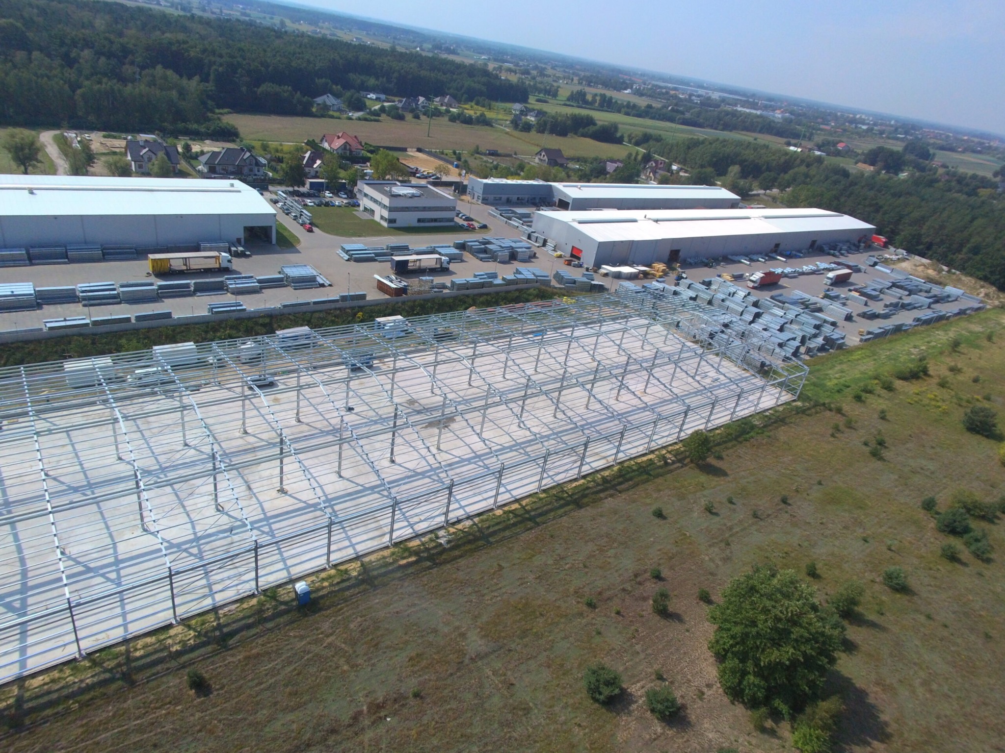 Szkielet konstrukcji stalowej hali w trakcie budowy, widok z góry, z widocznymi elementami konstrukcyjnymi, placem budowy i budynkami w tle.