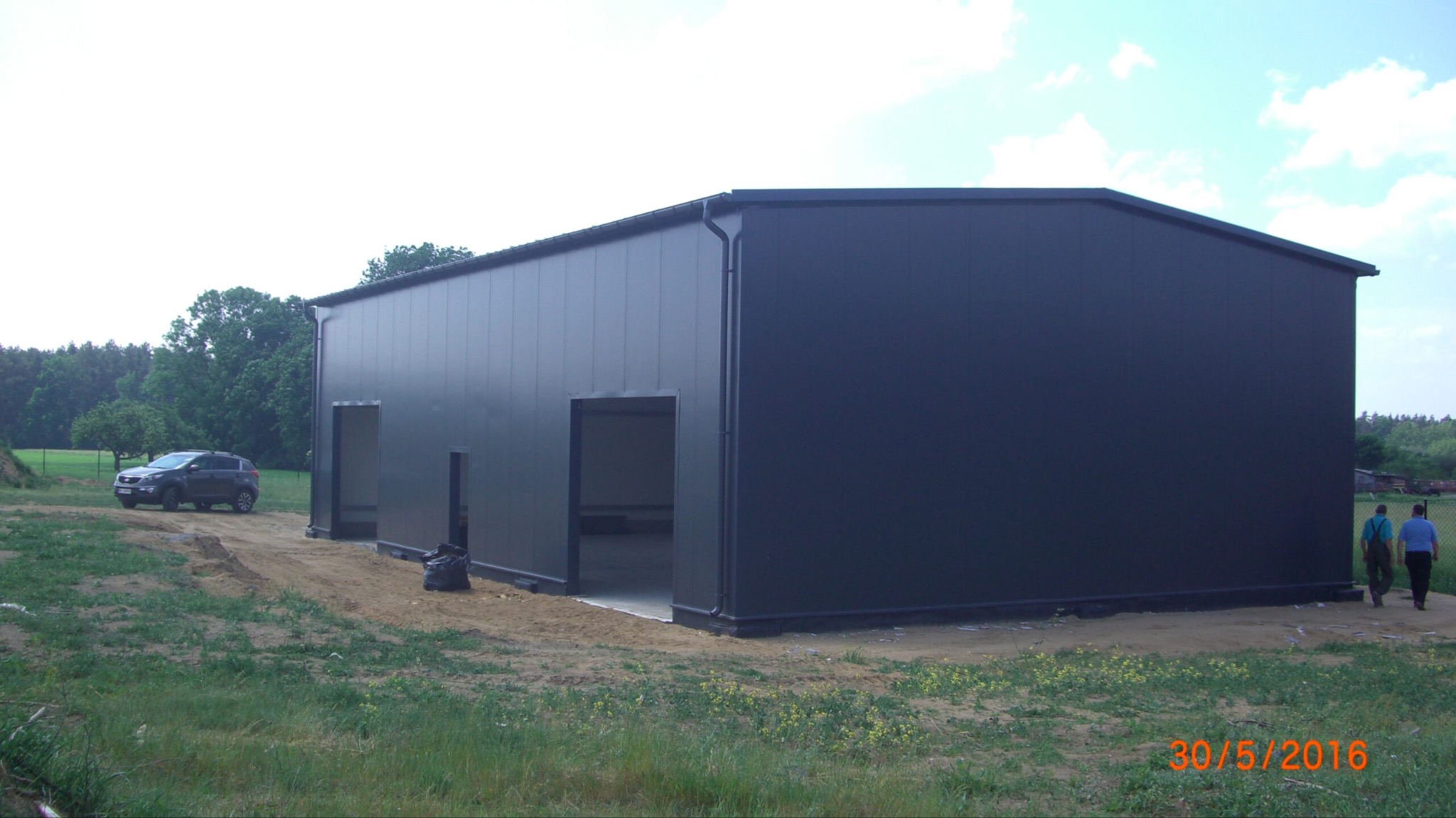 Gotowa hala z płyt warstwowych w kolorze grafitowym, widoczny samochód osobowy w tle i dwie osoby w oddali.