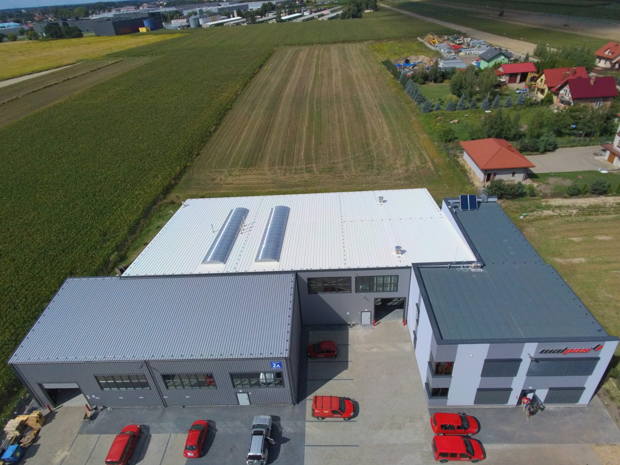 Zdjęcie z drona przedstawiające nowoczesną halę produkcyjną z szarej blachy trapezowej z oknami i bramami, z białym dachem z świetlikami dachowymi, otoczoną polami i zabudową jednorodzinną,...
