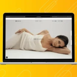 Laptop wyświetla stronę internetową z modelką prezentującą nową kolekcję ubrań na białym tle. Widoczny pasek adresu z nazwą domeny i logo firmy w lewym górnym rogu. Uwagę przykuwa minimalistyczny...