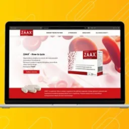 Laptop wyświetla stronę internetową suplementu diety ZAAX z czerwonym motywem przewodnim, prezentującą opakowanie produktu, kapsułki i hasło 'ZAAX - Krew to życie'.