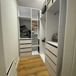 Biała garderoba wnękowa z szufladami, półkami i drążkami, wypełniona pudełkami, ubraniami i akcesoriami, oświetlona lampą sufitową